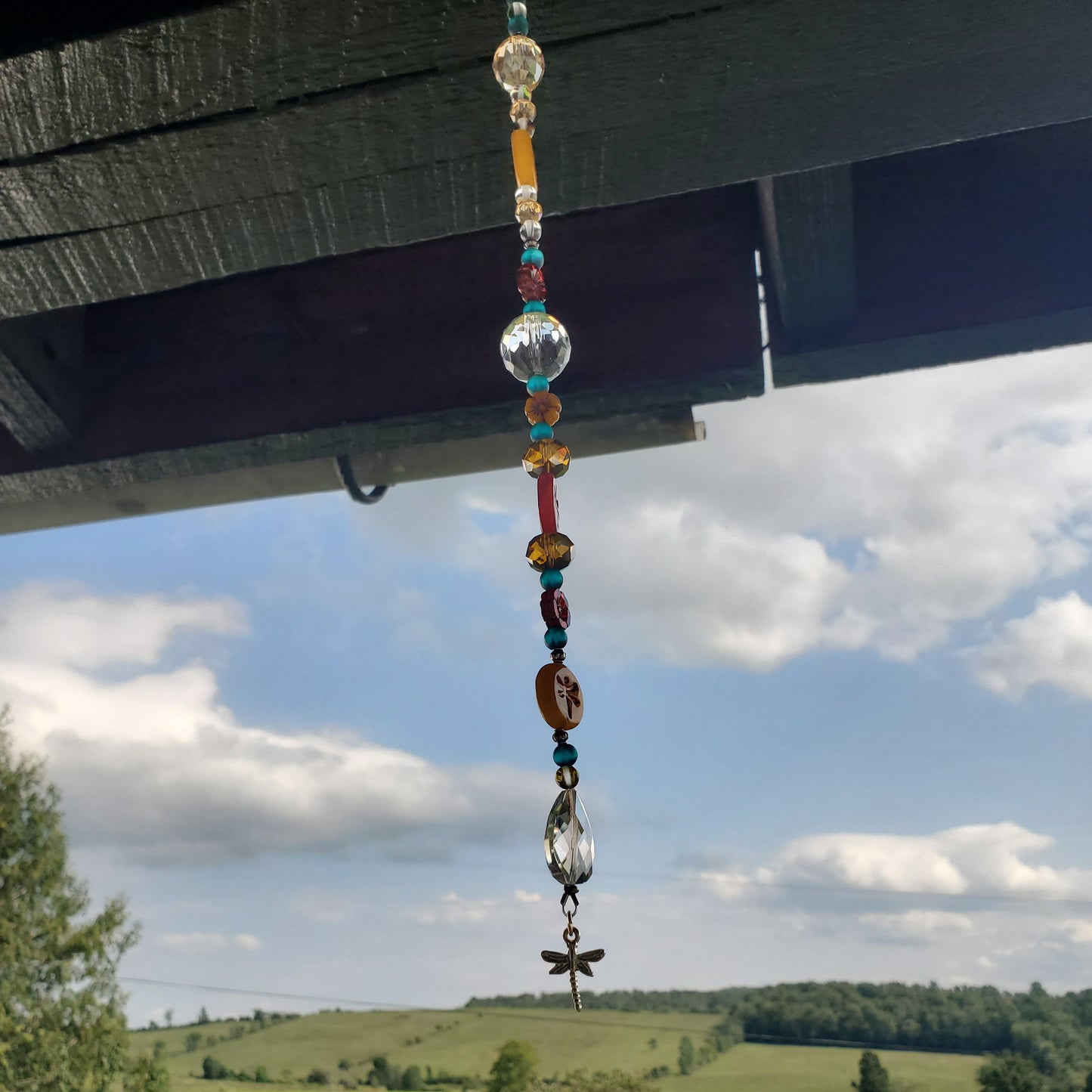 A colorful beaded suncatcher hangs from a dark wooden porch overhang, with a scenic green field and blue sky in the background.
