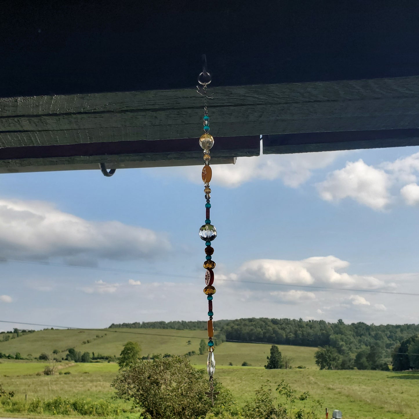 A colorful beaded hanging suncatcher is suspended from a dark bridge over a rural landscape with green fields, trees, and a blue sky with clouds.