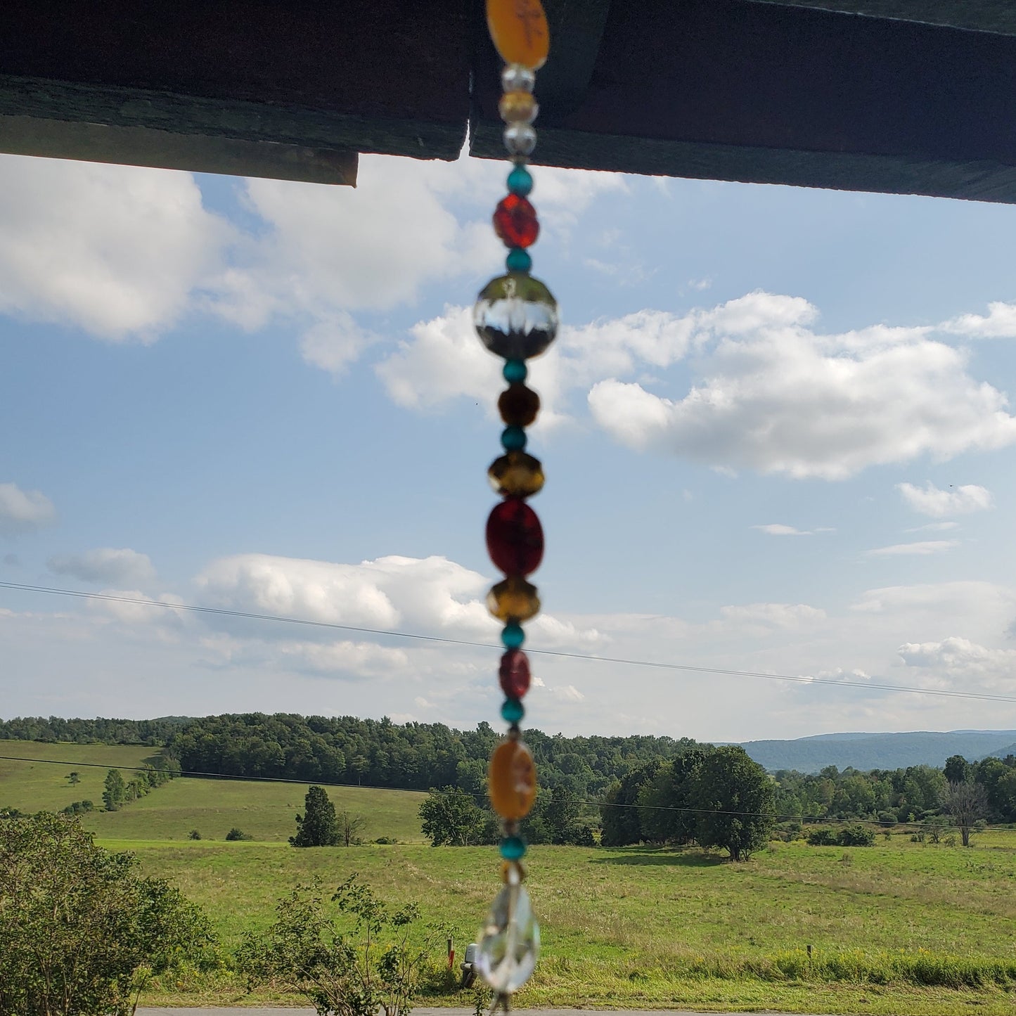 A vertical glass bead suncatcher hangs from a dark wooden eave, set against a scenic outdoor background of a green valley, trees, and a blue sky with clouds.