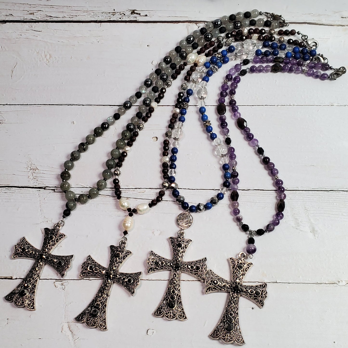Four beaded rosary necklaces with ornate silver-toned cross pendants are arranged on a white wooden surface. The necklaces feature beads in black, gray, blue, and purple, with the silver crosses hanging below.