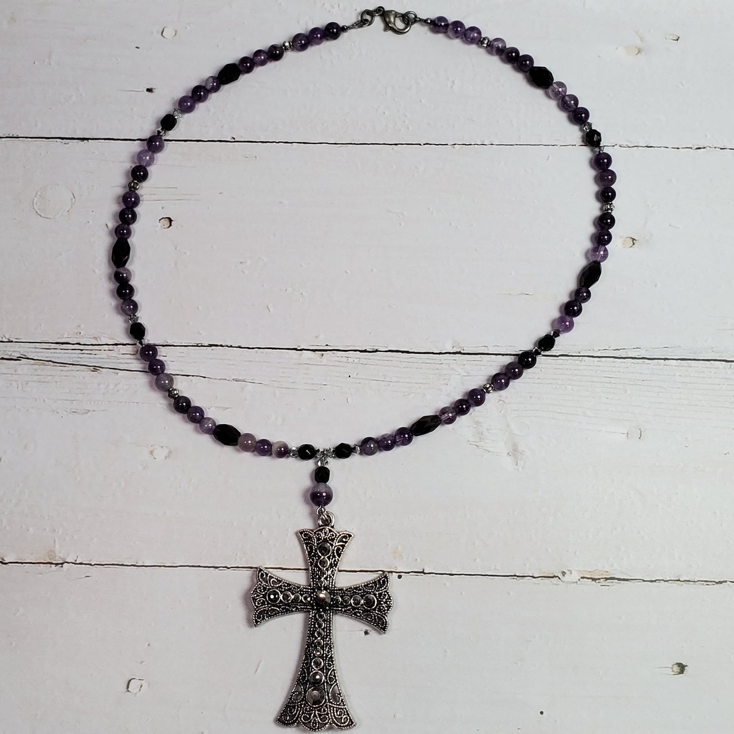 A beaded rosary necklace with dark purple beads and a silver-toned cross pendant, displayed on a white wooden surface.