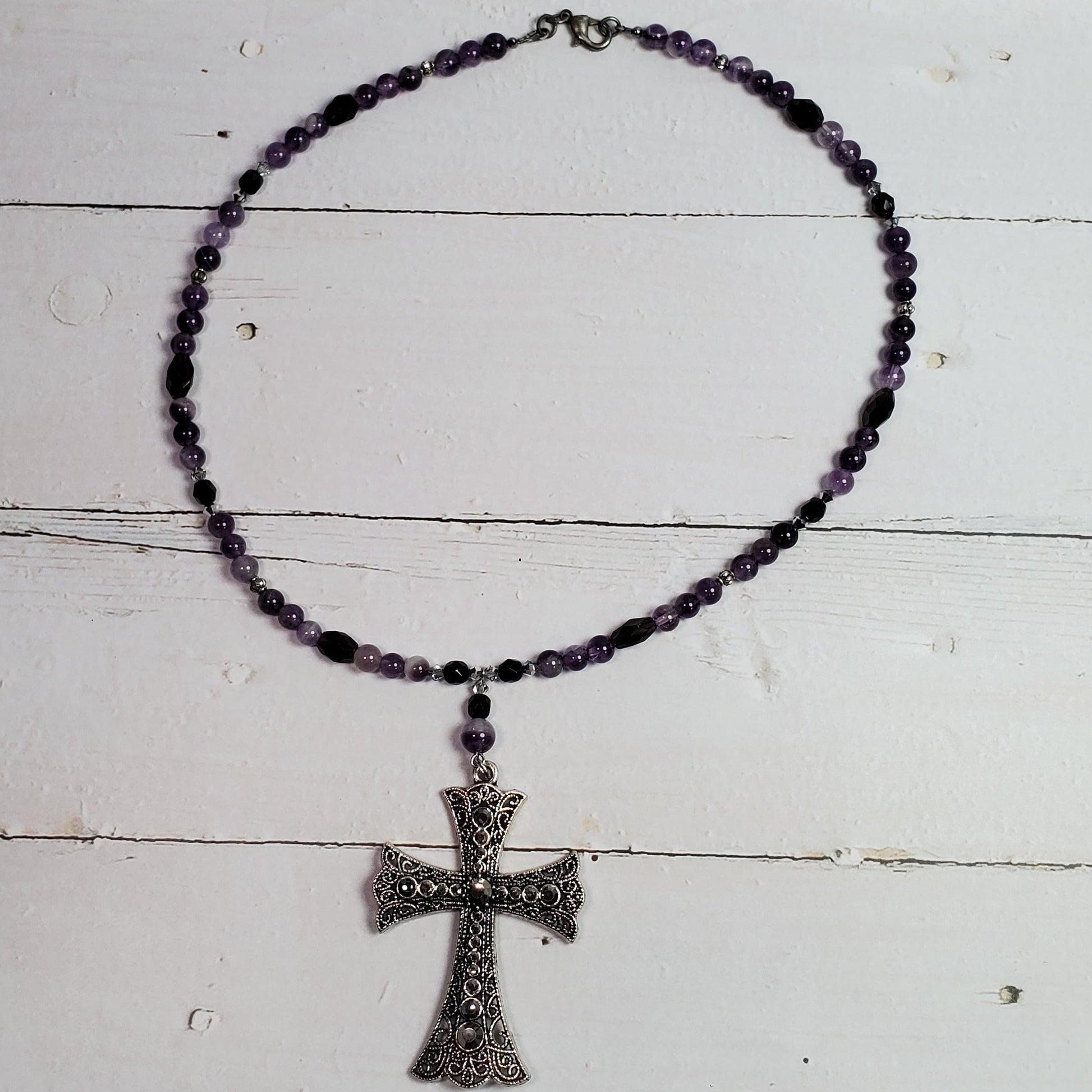 A beaded rosary necklace with dark purple beads and a silver-toned cross pendant, displayed on a white wooden surface.