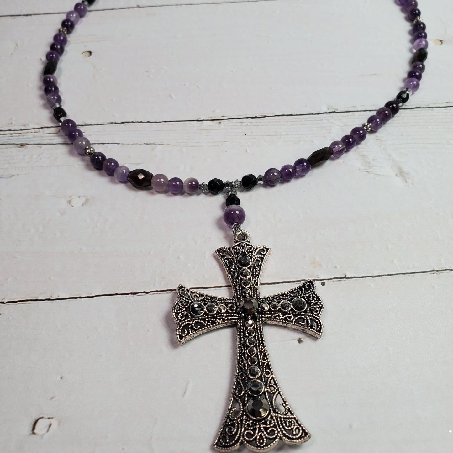 A beaded necklace with purple and black beads and a large ornate silver cross pendant, displayed on a white wooden surface.