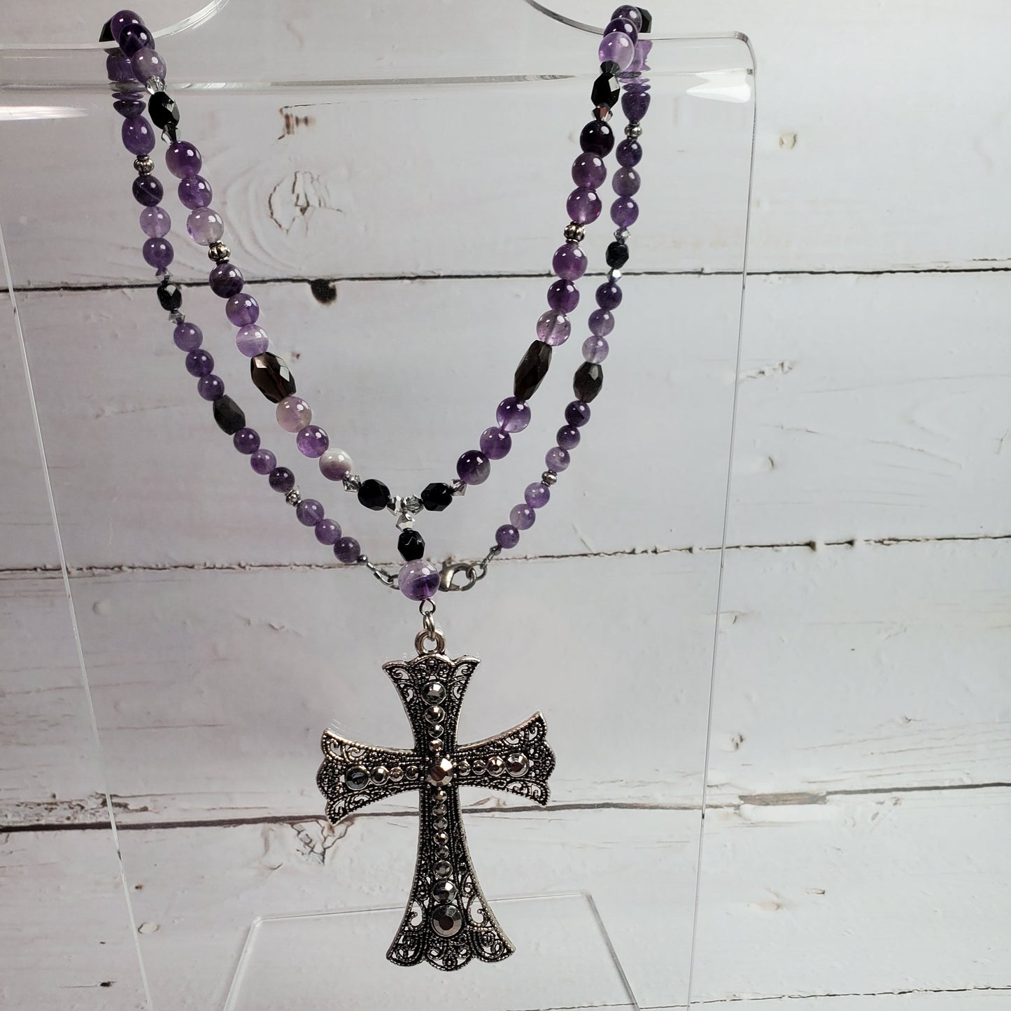 A beaded necklace with purple and black beads and a silver filigree cross pendant, displayed on a clear acrylic stand against a white wood plank background.