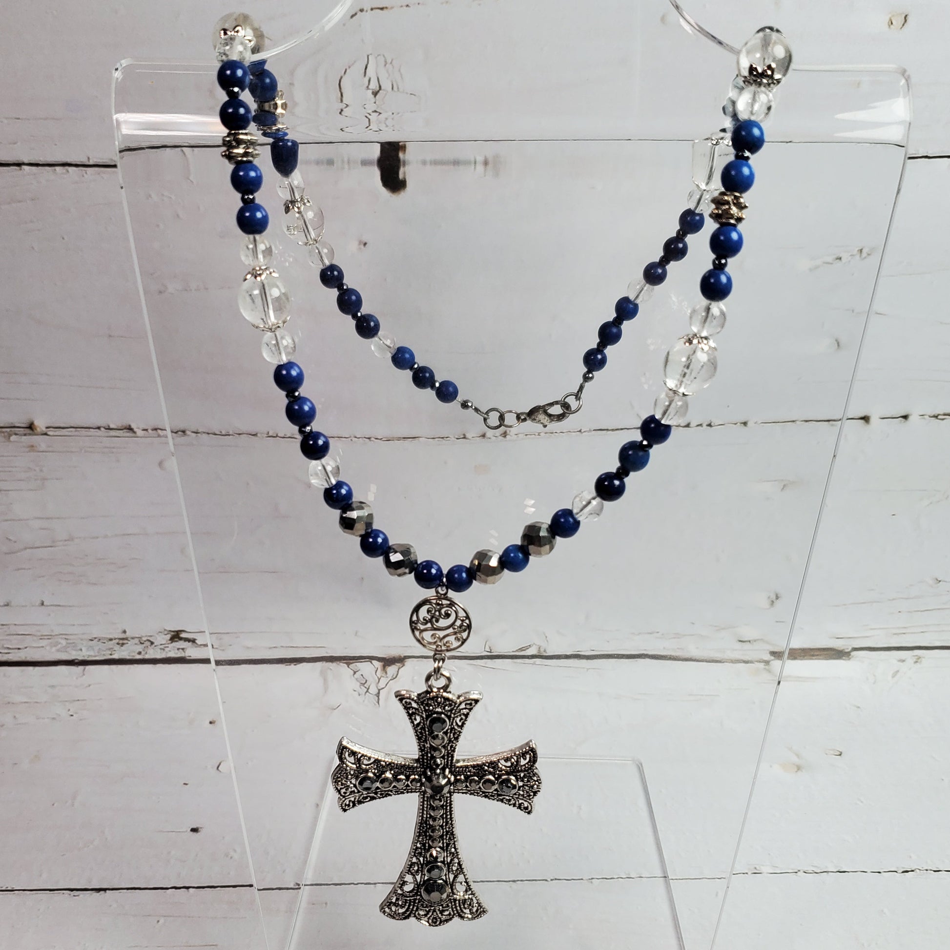 A rosary-style necklace with dark blue beads, clear faceted beads, and a silver-toned ornate cross pendant, displayed on a clear acrylic stand against a white wood plank background.