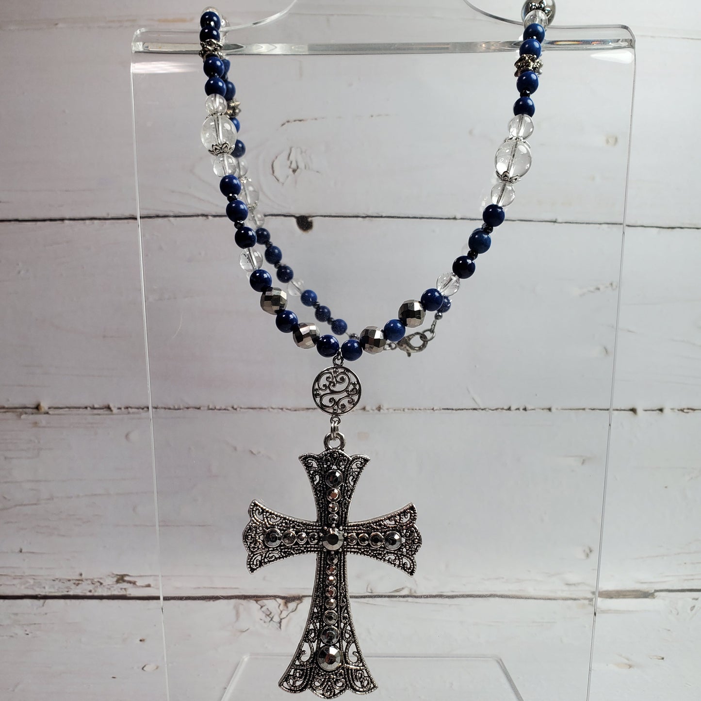 A beaded rosary necklace with dark blue and silver beads, featuring an ornate silver-toned cross pendant, displayed on a clear acrylic stand against a white wood plank background.