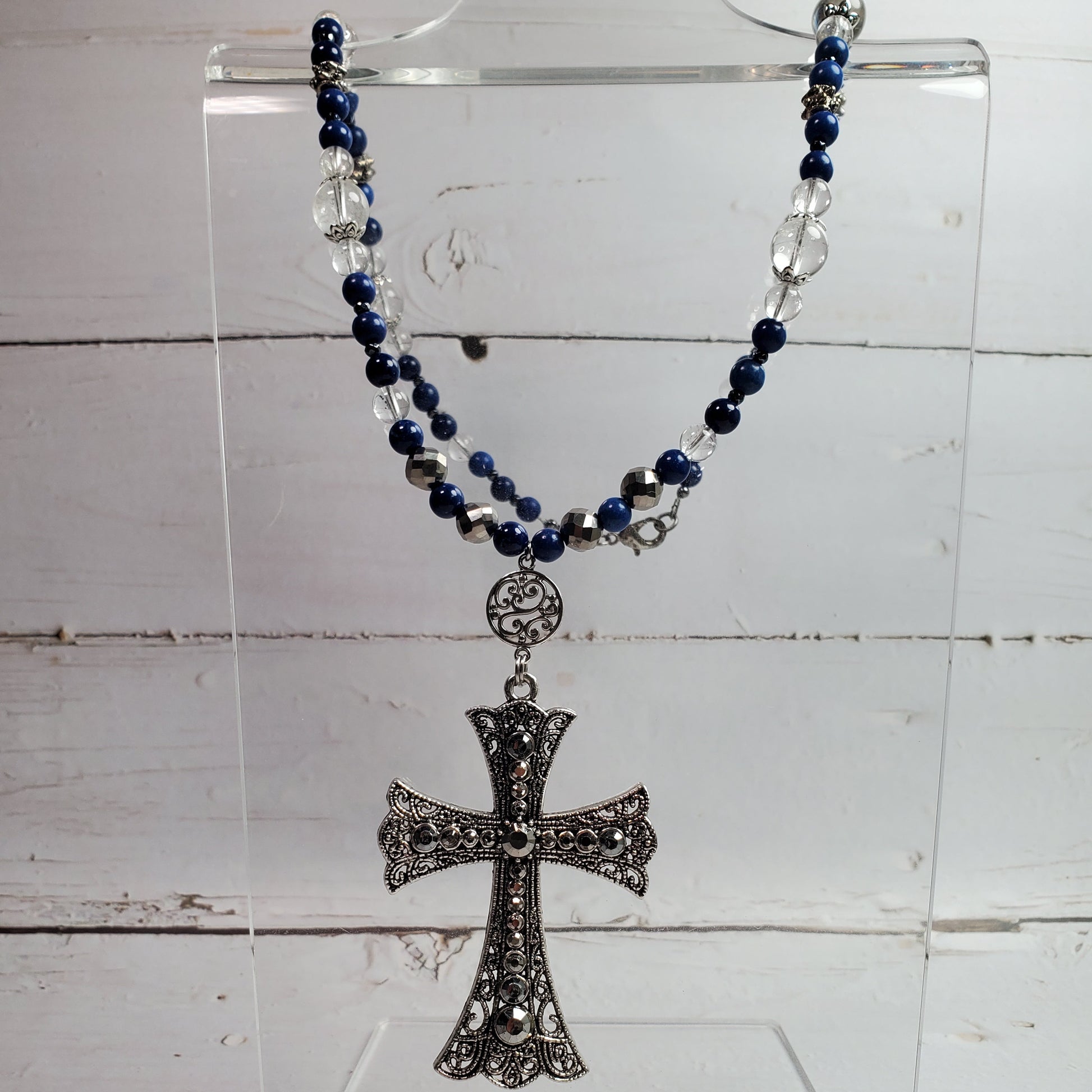 A beaded rosary necklace with dark blue and silver beads, featuring an ornate silver-toned cross pendant, displayed on a clear acrylic stand against a white wood plank background.
