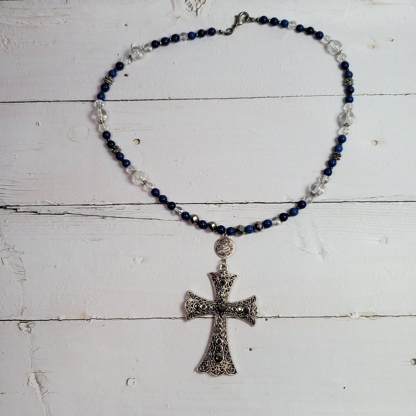 A beaded necklace with dark blue and clear faceted beads, featuring an ornate silver-toned cross pendant, displayed on a white wooden plank surface.