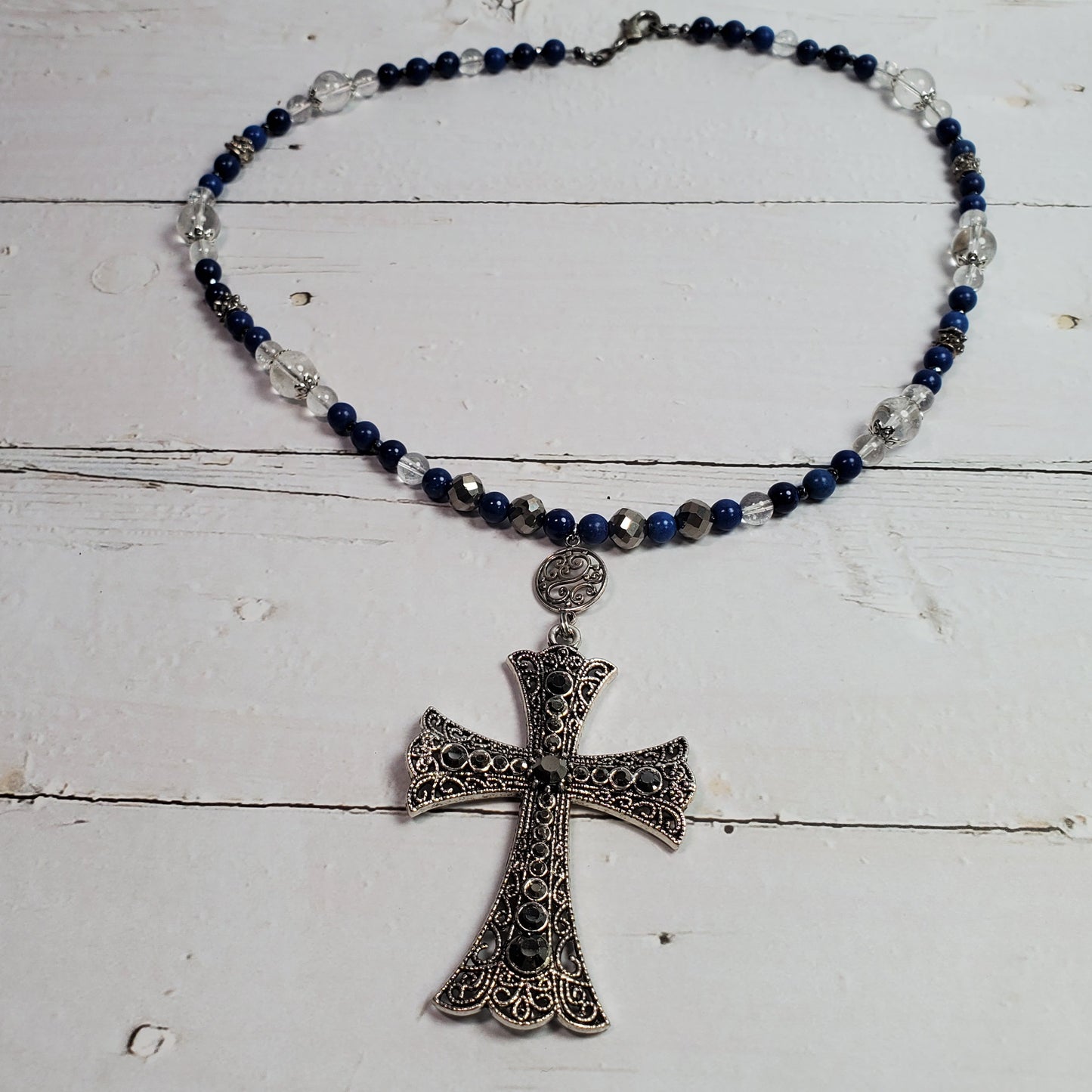 A beaded necklace with dark blue and clear faceted beads, featuring an ornate silver filigree cross pendant, displayed on a white wooden surface.