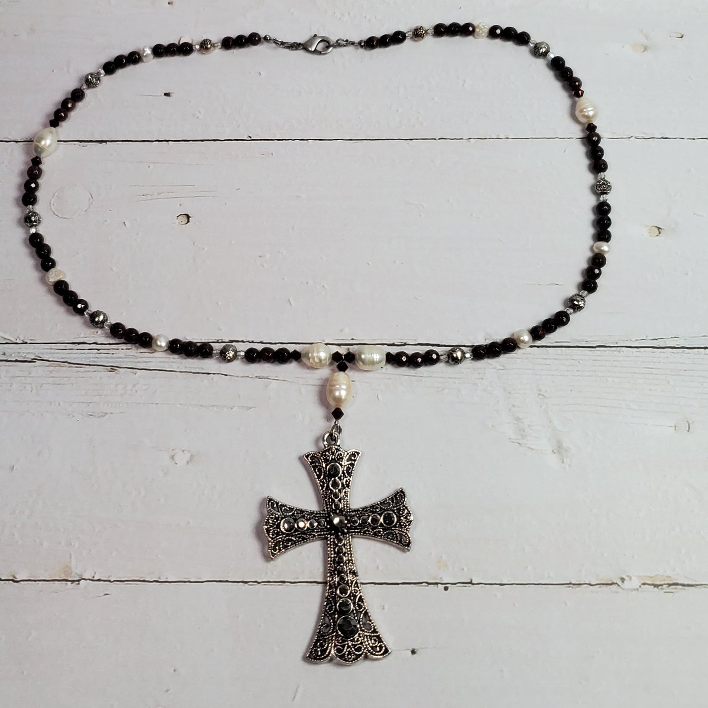 A beaded necklace with black and white beads and a silver-toned cross pendant, displayed on a white wooden plank background.