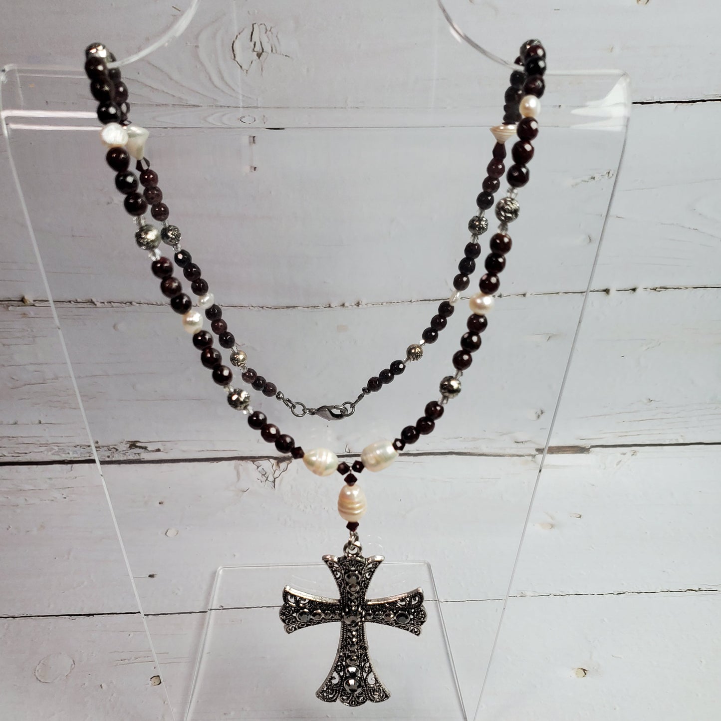 A beaded necklace with dark red beads, small white pearl-like accents, and a textured silver-toned cross pendant, displayed on a clear acrylic stand against a white wood background.