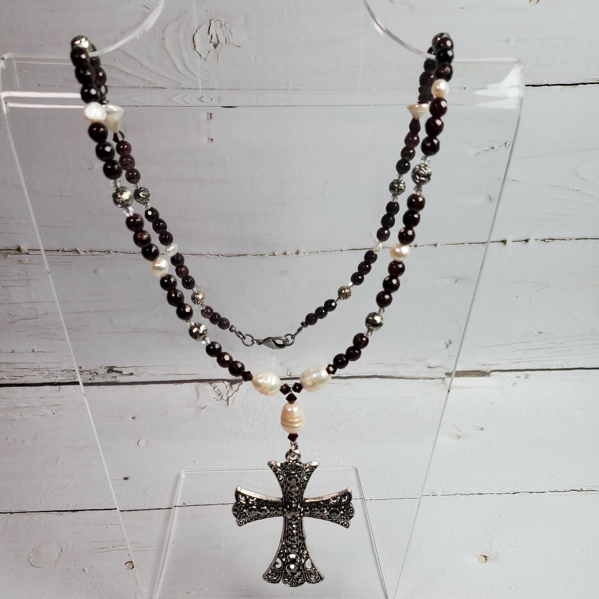 A beaded necklace with dark red beads, small white pearl-like accents, and a textured silver-toned cross pendant, displayed on a clear acrylic stand against a white wood background.