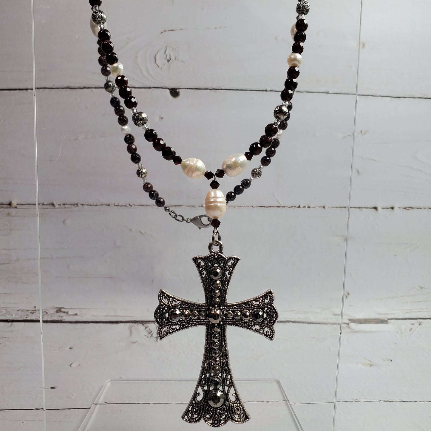 A rosary necklace with black and white beads and a detailed filigree cross pendant, displayed on a clear acrylic stand against a white wooden background.