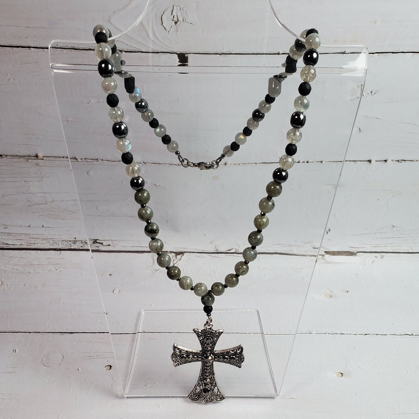 A beaded necklace with dark gray, black, and light gray beads, featuring a textured silver-toned cross pendant, displayed on a clear acrylic stand against a white wood plank background.