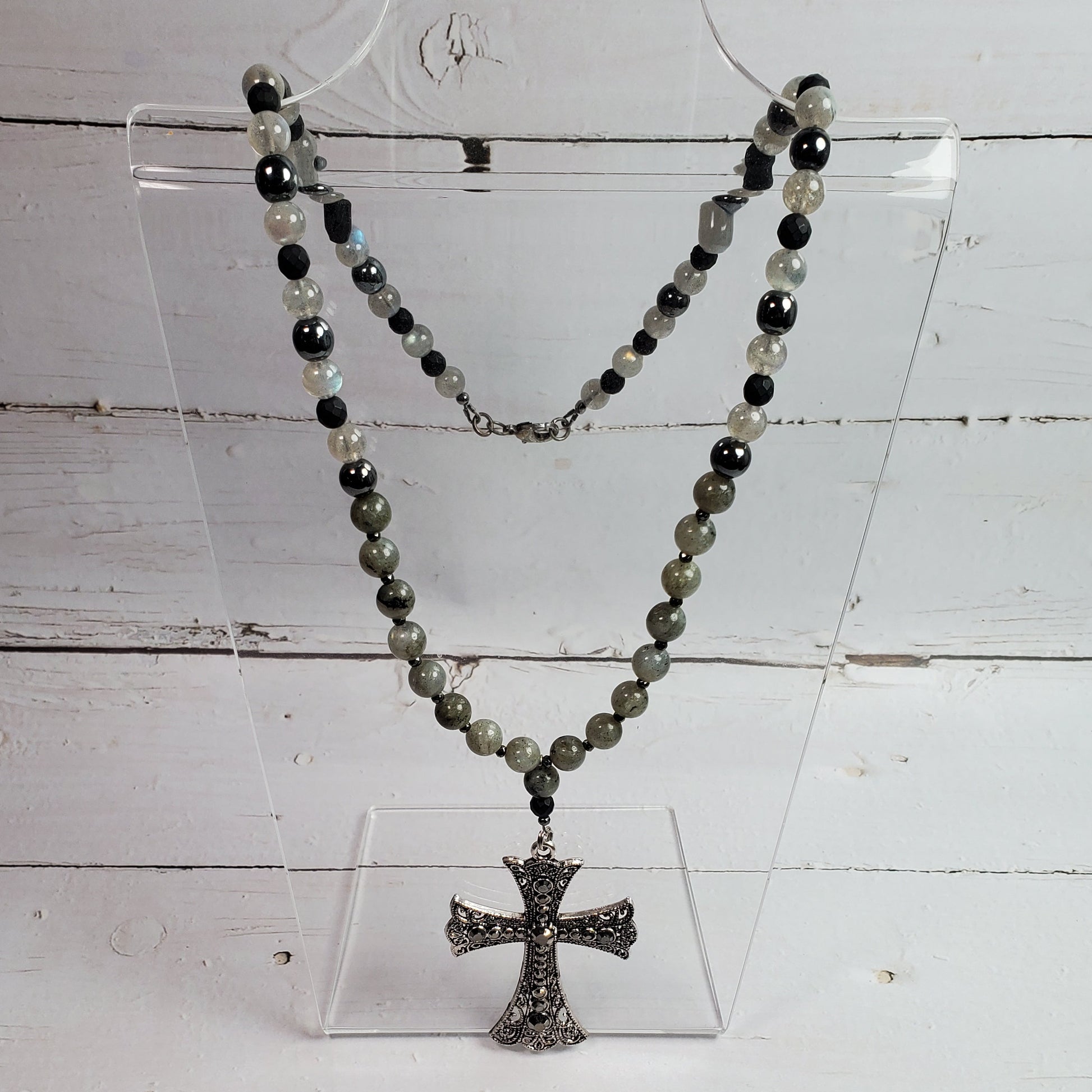 A beaded necklace with dark gray, black, and light gray beads, featuring a textured silver-toned cross pendant, displayed on a clear acrylic stand against a white wood plank background.