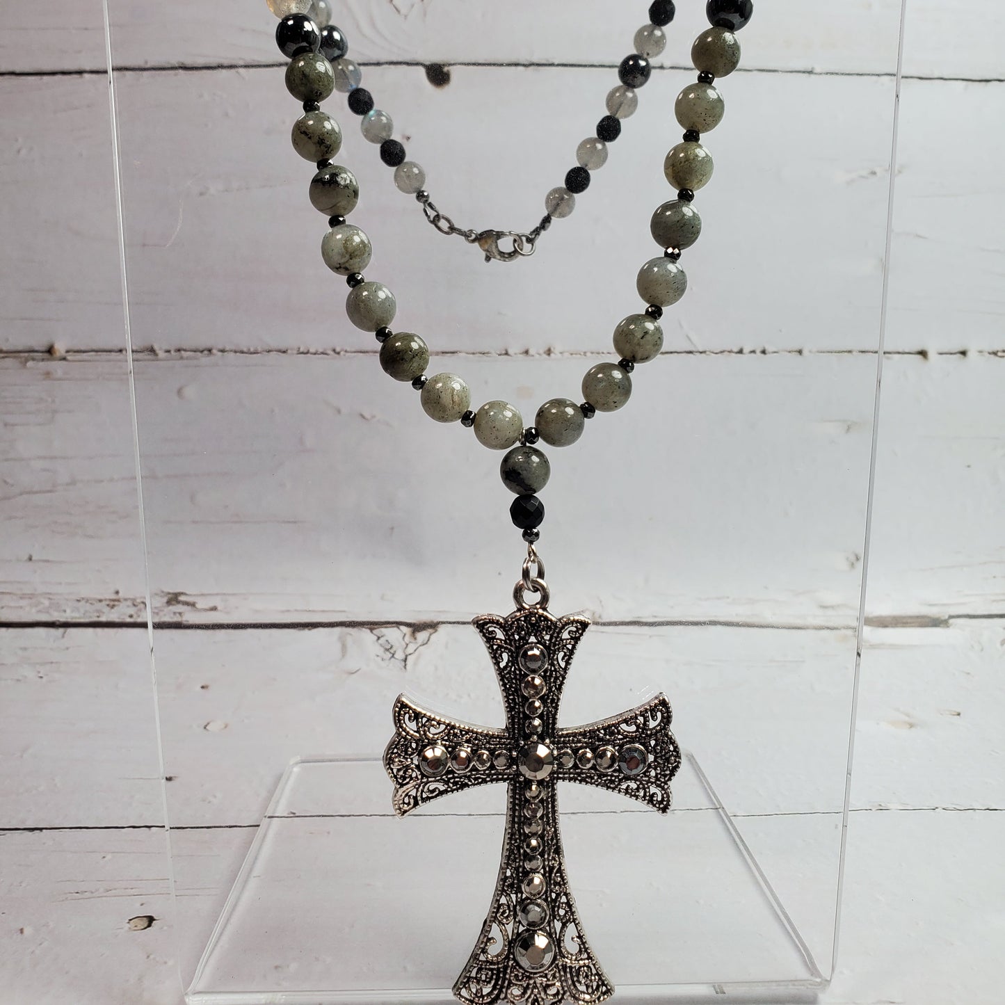 A beaded necklace with a filigree cross pendant, displayed on a clear acrylic stand against a white wood plank background. The beads are a mix of gray, black, and translucent white stones, and the pendant is an antiqued silver-tone metal.