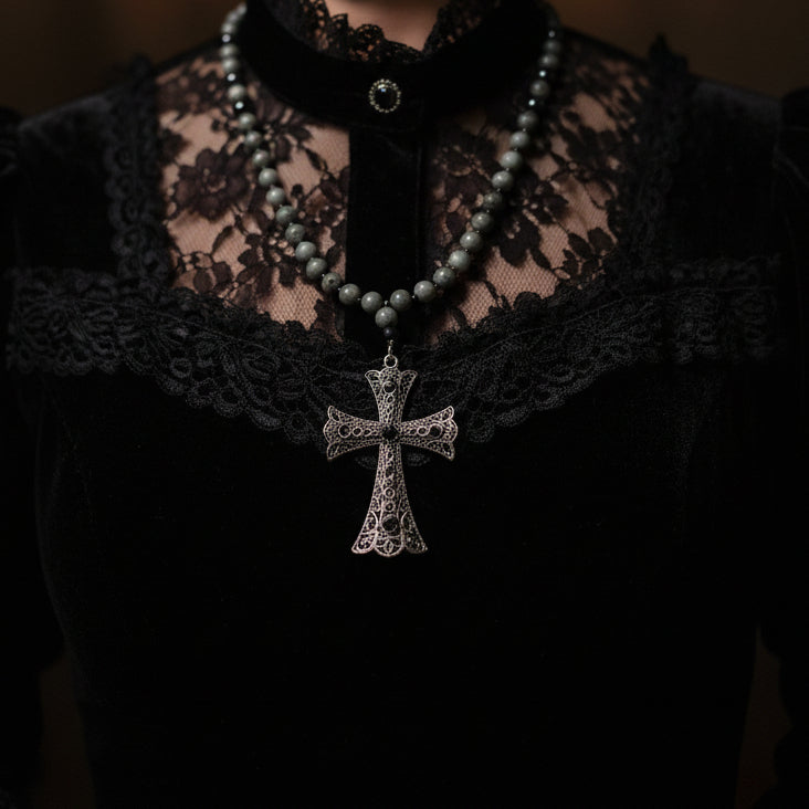 A beaded rosary necklace with dark gray beads and a textured silver-toned cross pendant, laid flat on a white wooden surface.
