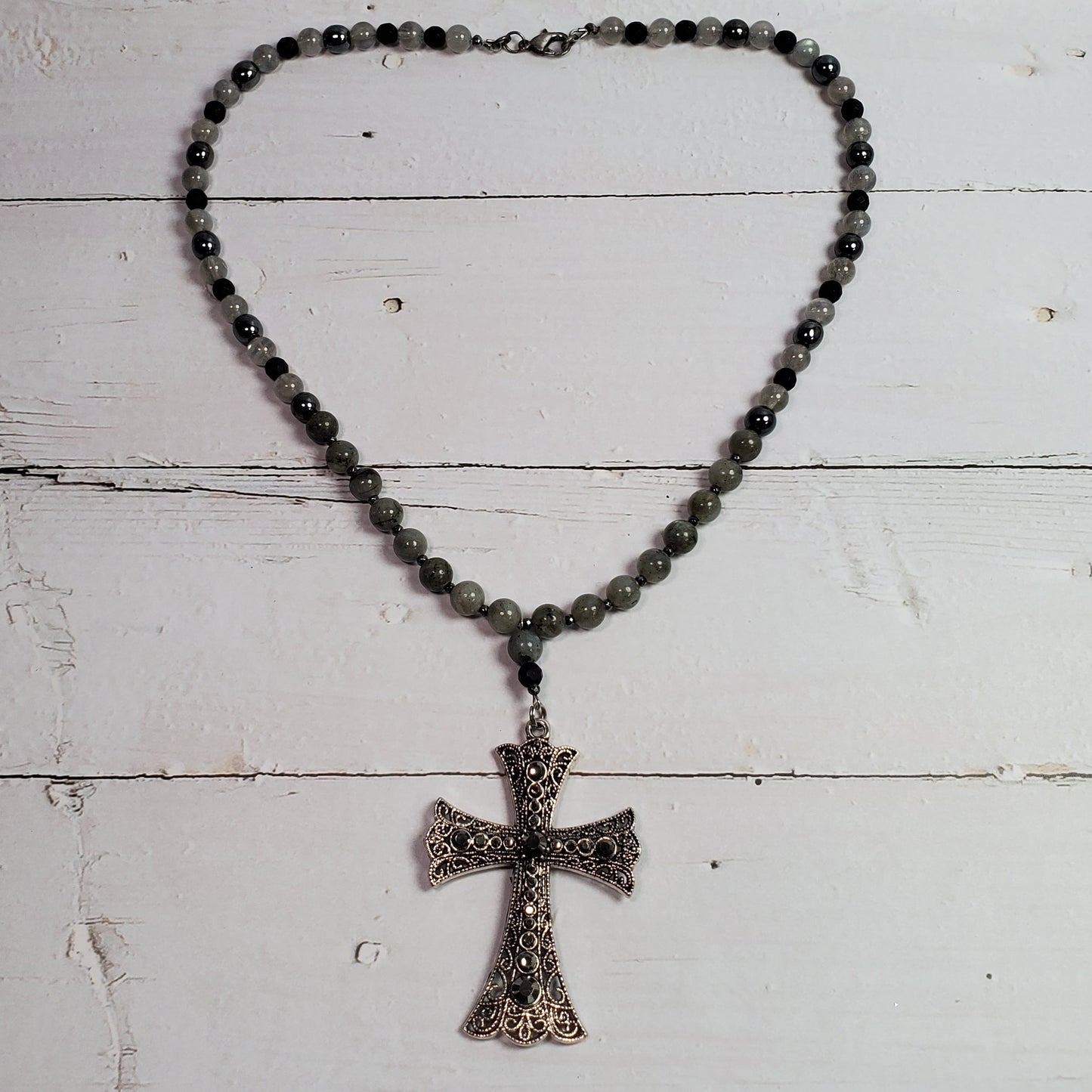 A beaded necklace with dark gray beads and a textured, antiqued bronze cross pendant, displayed on a white wooden plank surface.
