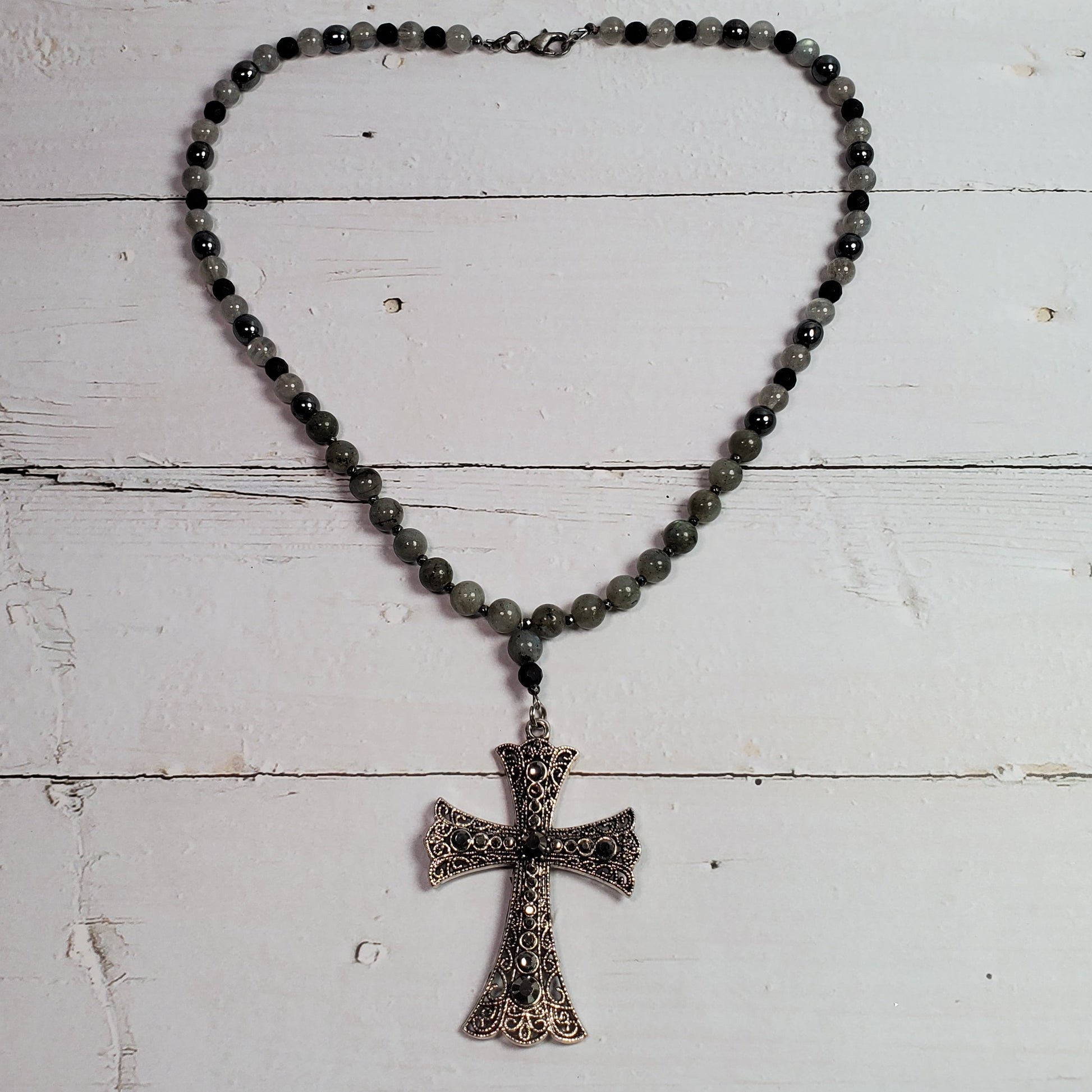 A beaded necklace with dark gray beads and a textured, antiqued bronze cross pendant, displayed on a white wooden plank surface.
