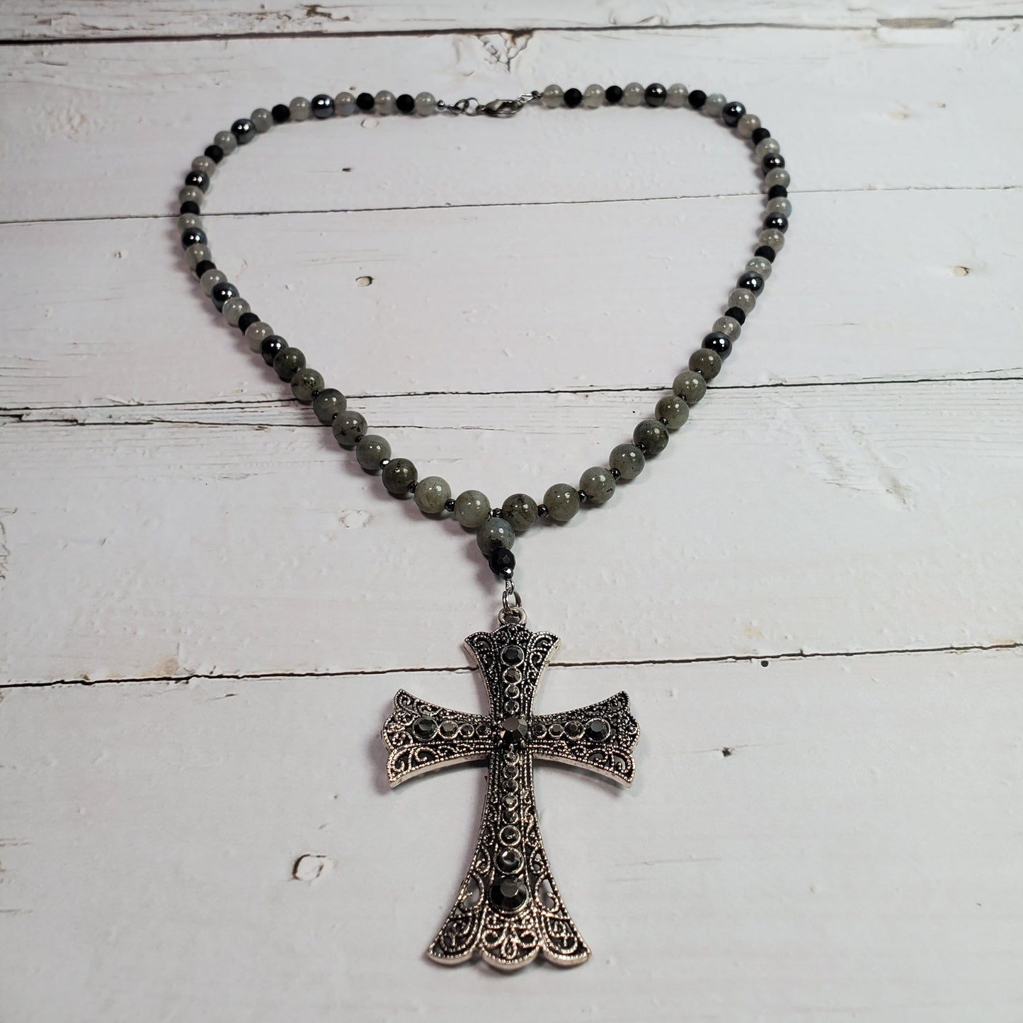 A beaded necklace with dark gray to black round beads and a silver-toned filigree cross pendant, displayed on a white wooden plank surface.