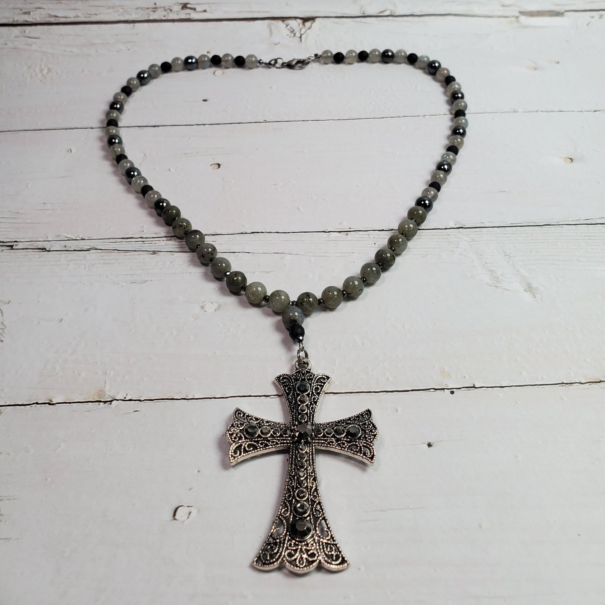 A beaded necklace with dark gray to black round beads and a silver-toned filigree cross pendant, displayed on a white wooden plank surface.