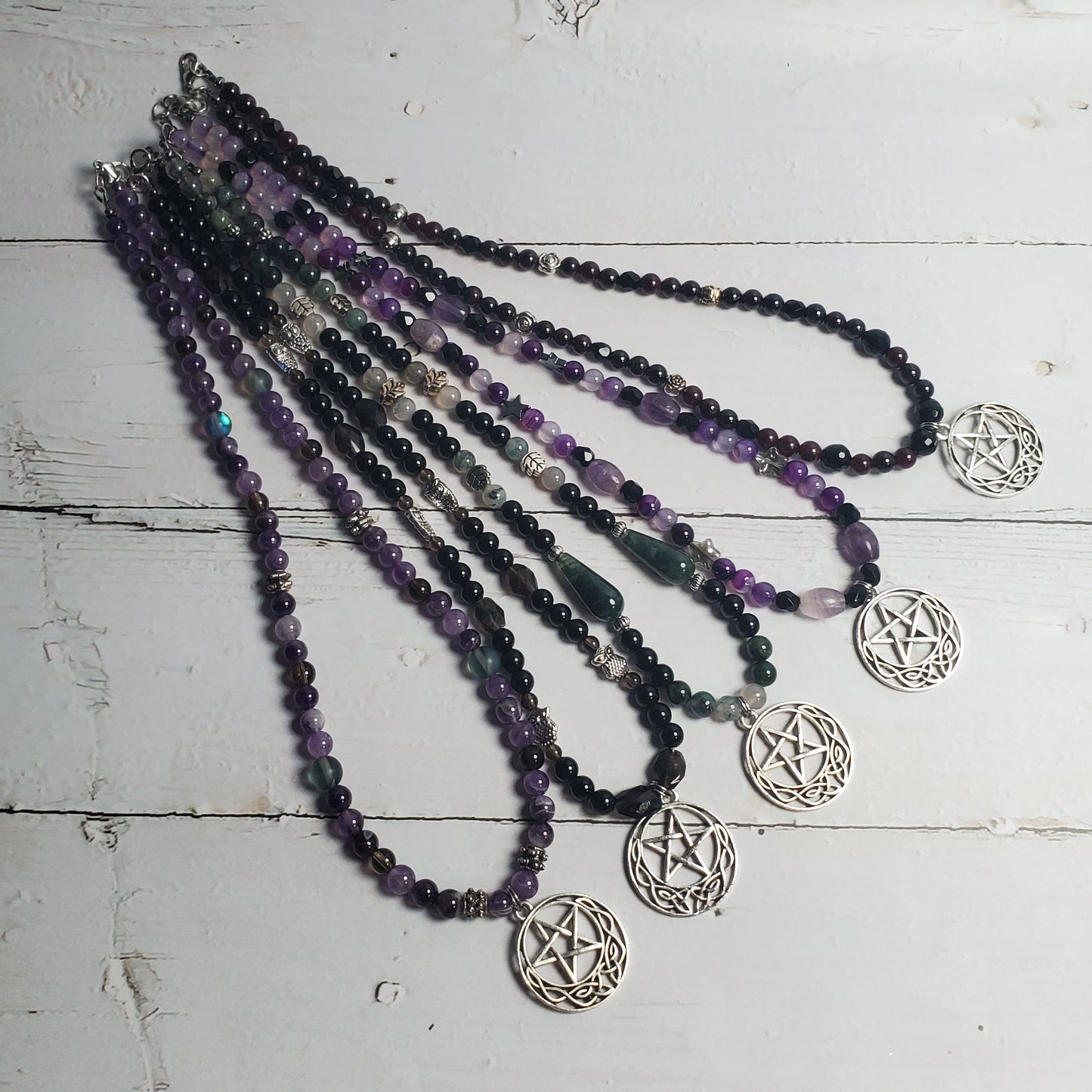 A set of five beaded necklaces with silver pentagram charms, arranged on a distressed white wooden surface. The necklaces feature beads in black, purple, and clear/white.