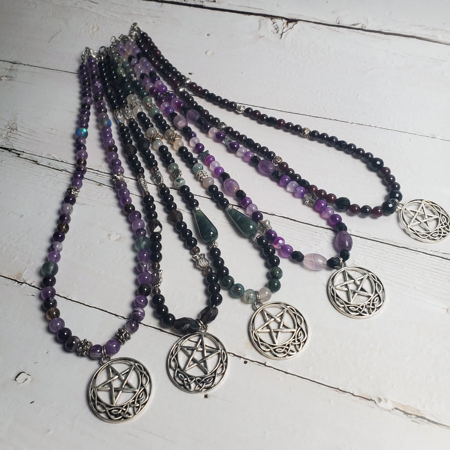 Five beaded necklaces with silver pentagram pendants arranged on a white wooden surface. The necklaces feature beads in purple, black, green, and clear quartz.