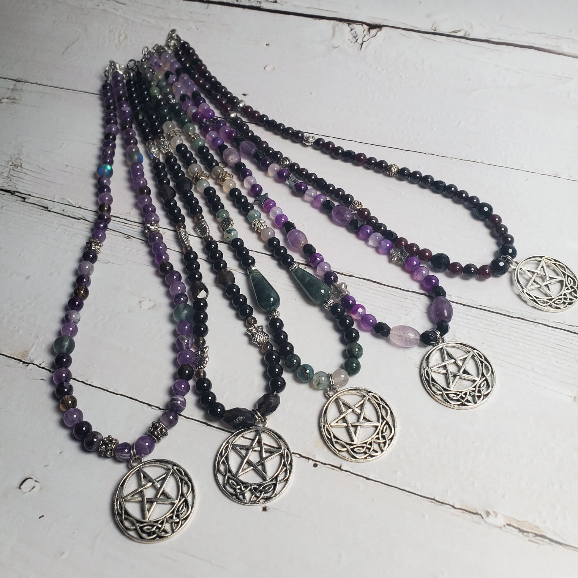 Five beaded necklaces with silver pentagram pendants arranged on a white wooden surface. The necklaces feature beads in purple, black, green, and clear quartz.