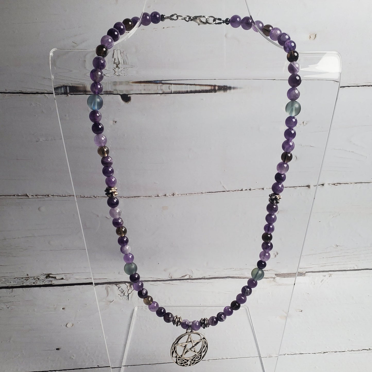 A beaded necklace with purple, gray, and light blue beads, featuring a silver-toned circular pendant with a star design, displayed on a clear acrylic stand against a white wooden background.