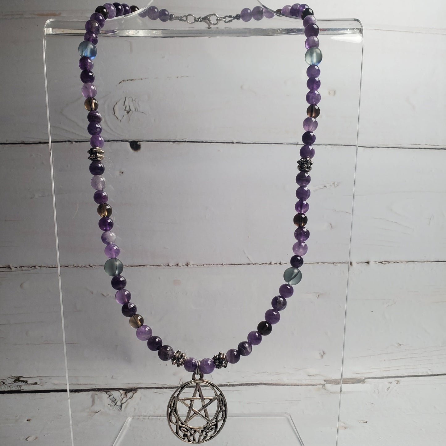 A beaded necklace with purple, gray, and blue beads, featuring a silver-toned pentagram sphere pendant, displayed on a clear acrylic stand against a white wood plank background.
