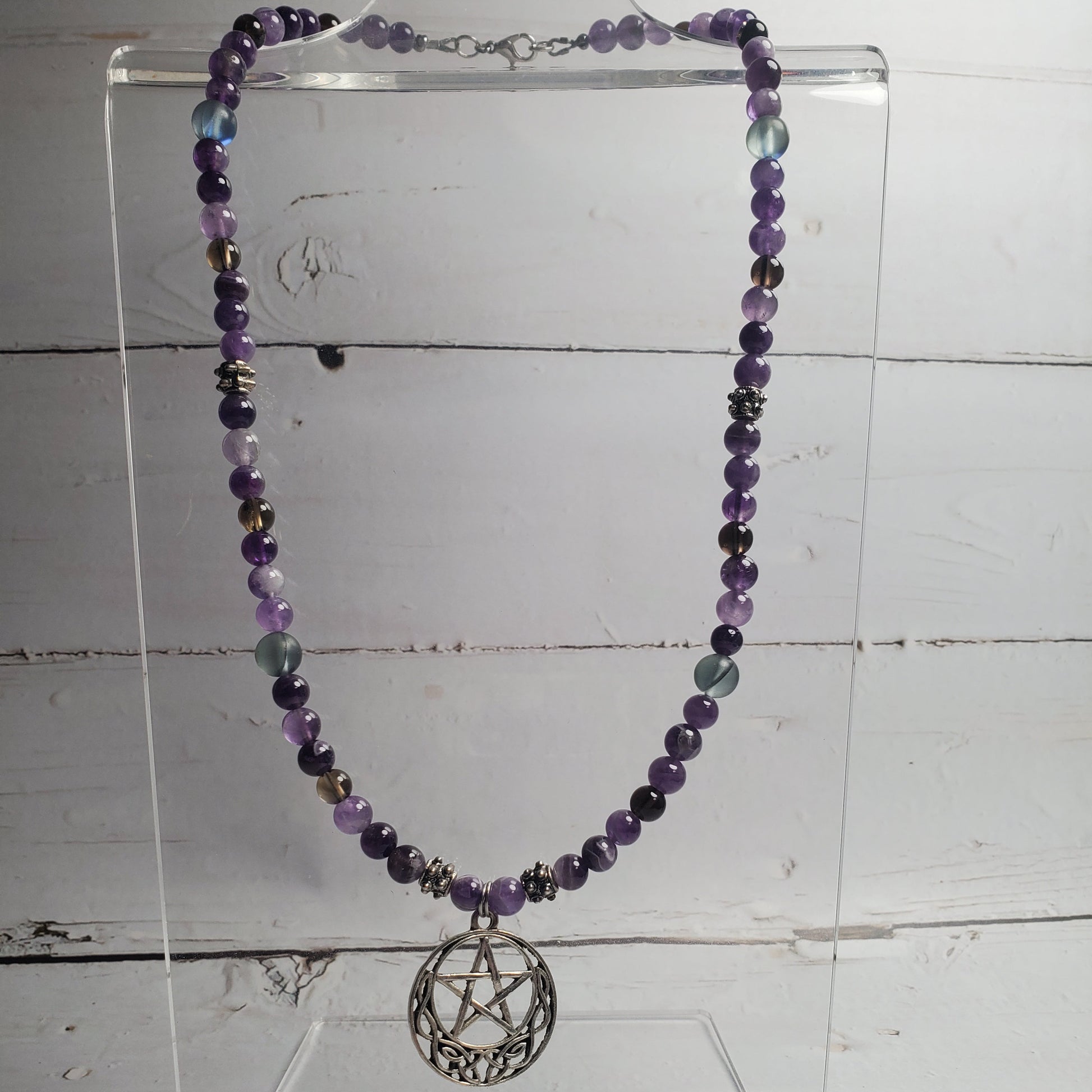 A beaded necklace with purple, gray, and blue beads, featuring a silver-toned pentagram sphere pendant, displayed on a clear acrylic stand against a white wood plank background.