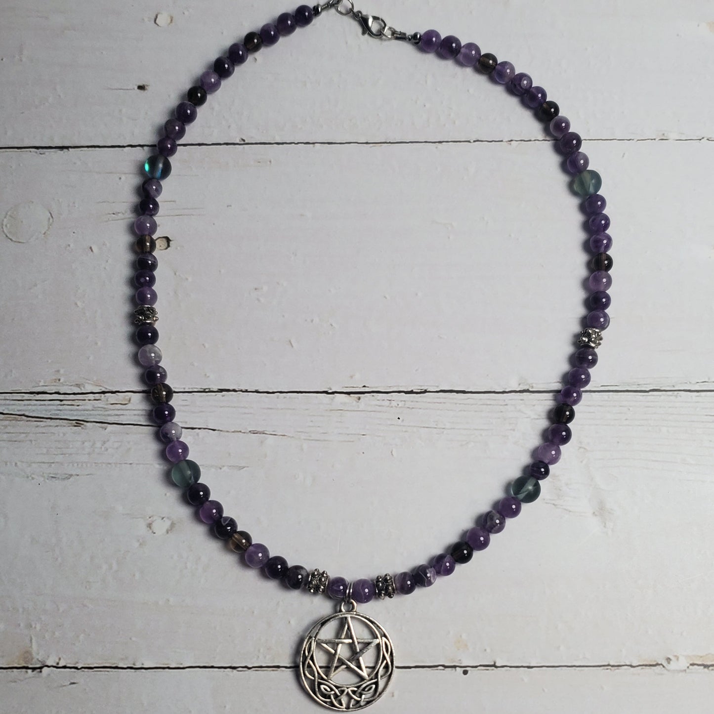 A beaded necklace with dark purple and green translucent beads, featuring a silver-toned pentagram pendant, displayed on a white wooden plank background.