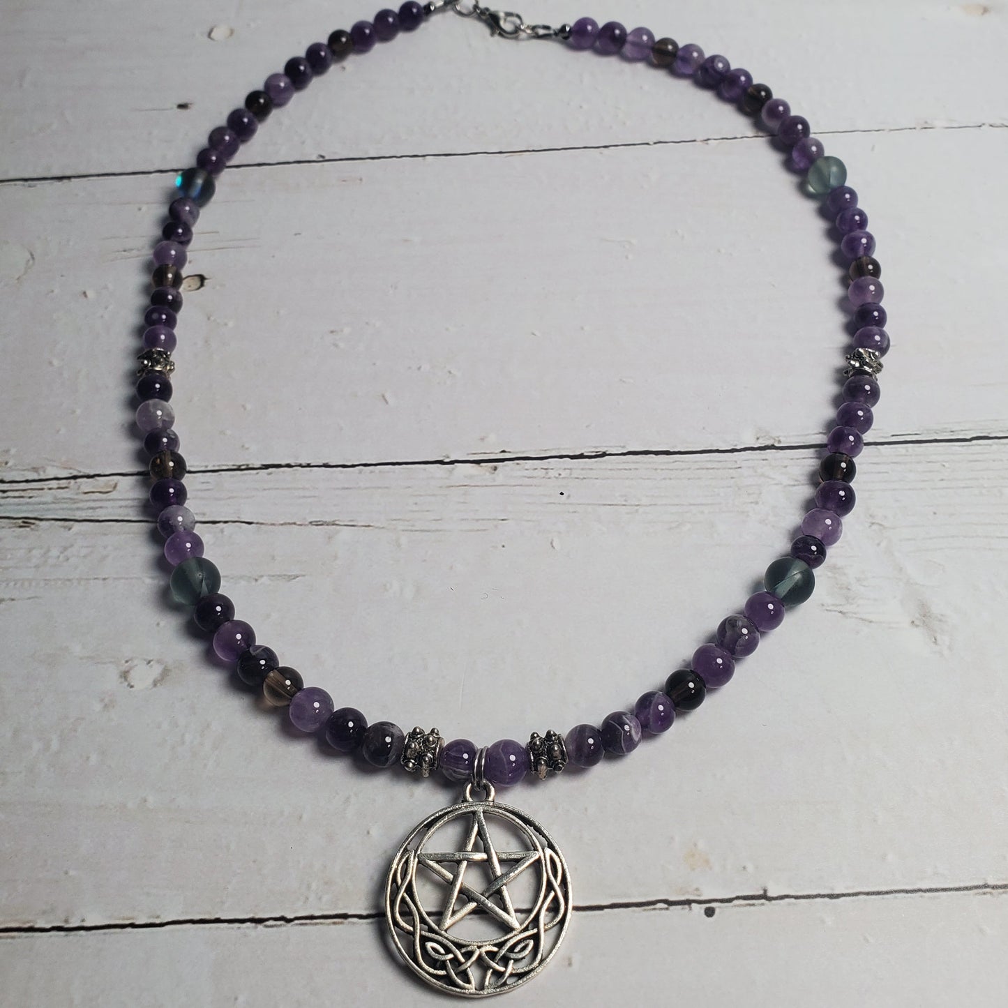 A beaded necklace with purple and black beads, featuring a silver-toned pentagram pendant, displayed on a white wooden surface.