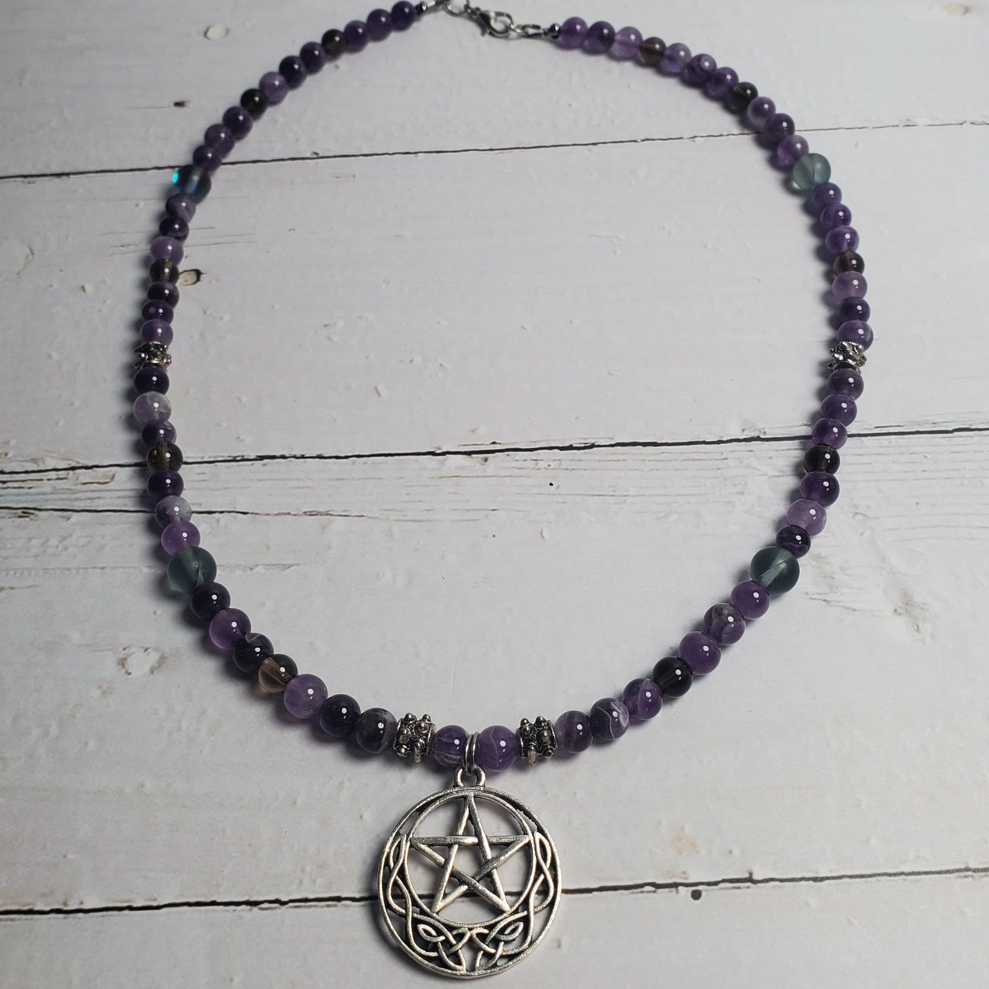 A beaded necklace with purple and black beads, featuring a silver-toned pentagram pendant, displayed on a white wooden surface.