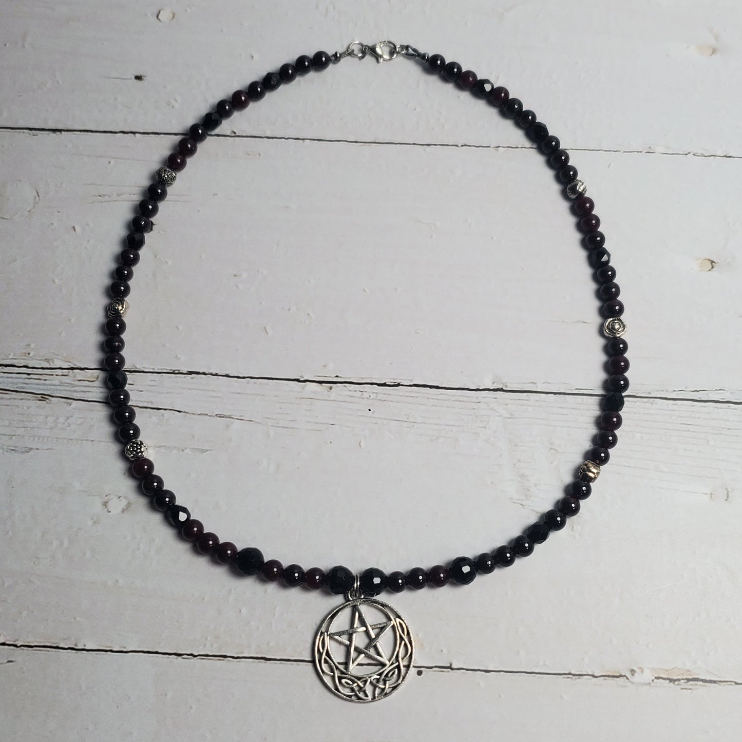 A beaded necklace with small dark beads and a silver-toned pentagram pendant, displayed on a distressed white wooden surface.