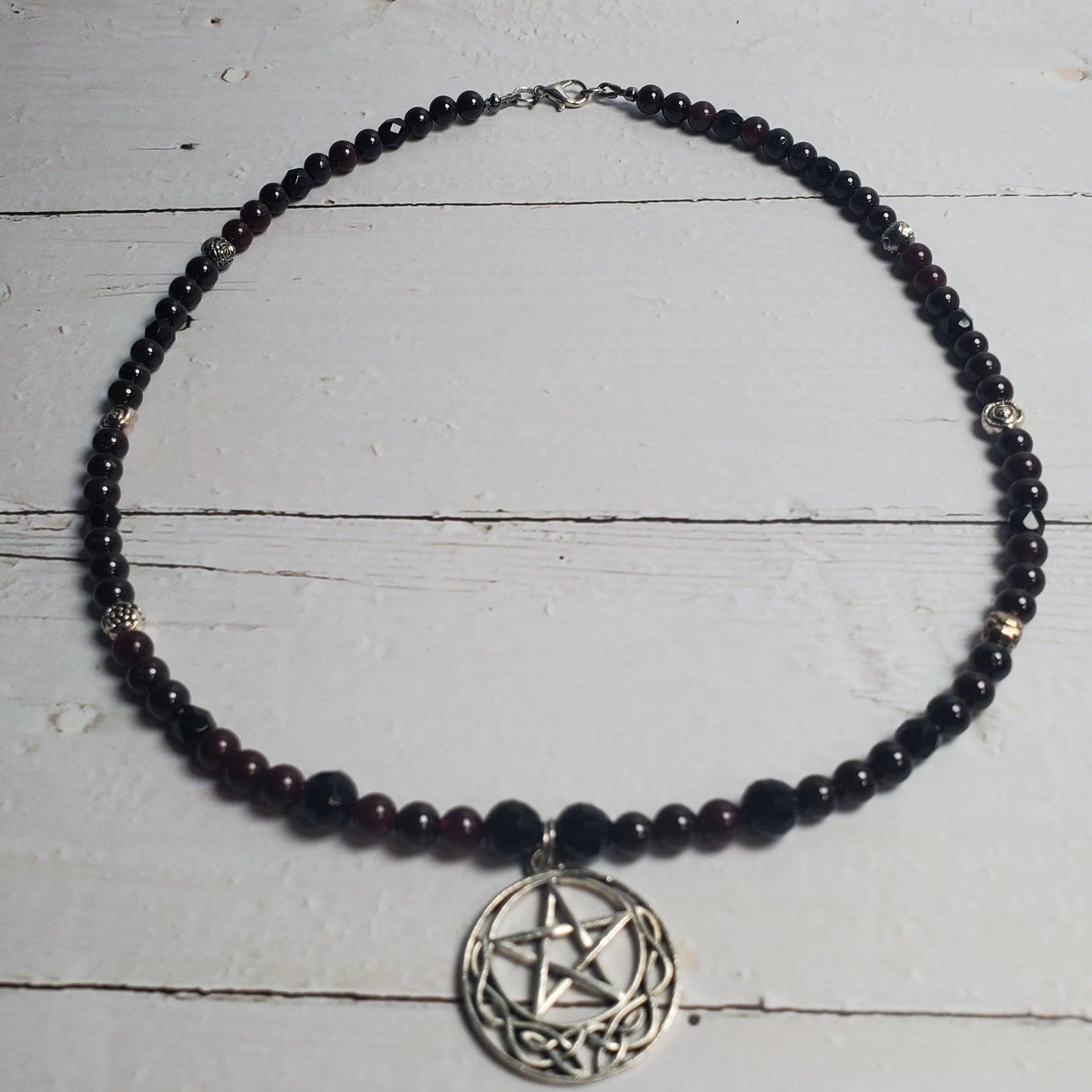 A beaded necklace with dark faceted beads and a silver-toned pentagram pendant, displayed on a distressed white wooden surface.