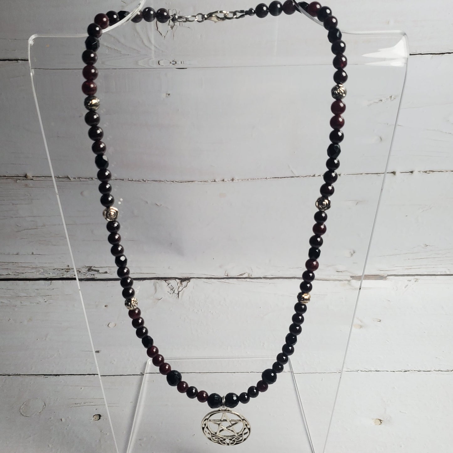 A beaded necklace with dark faceted beads and a silver-toned circular pendant, displayed on a clear acrylic stand against a white wood plank background.