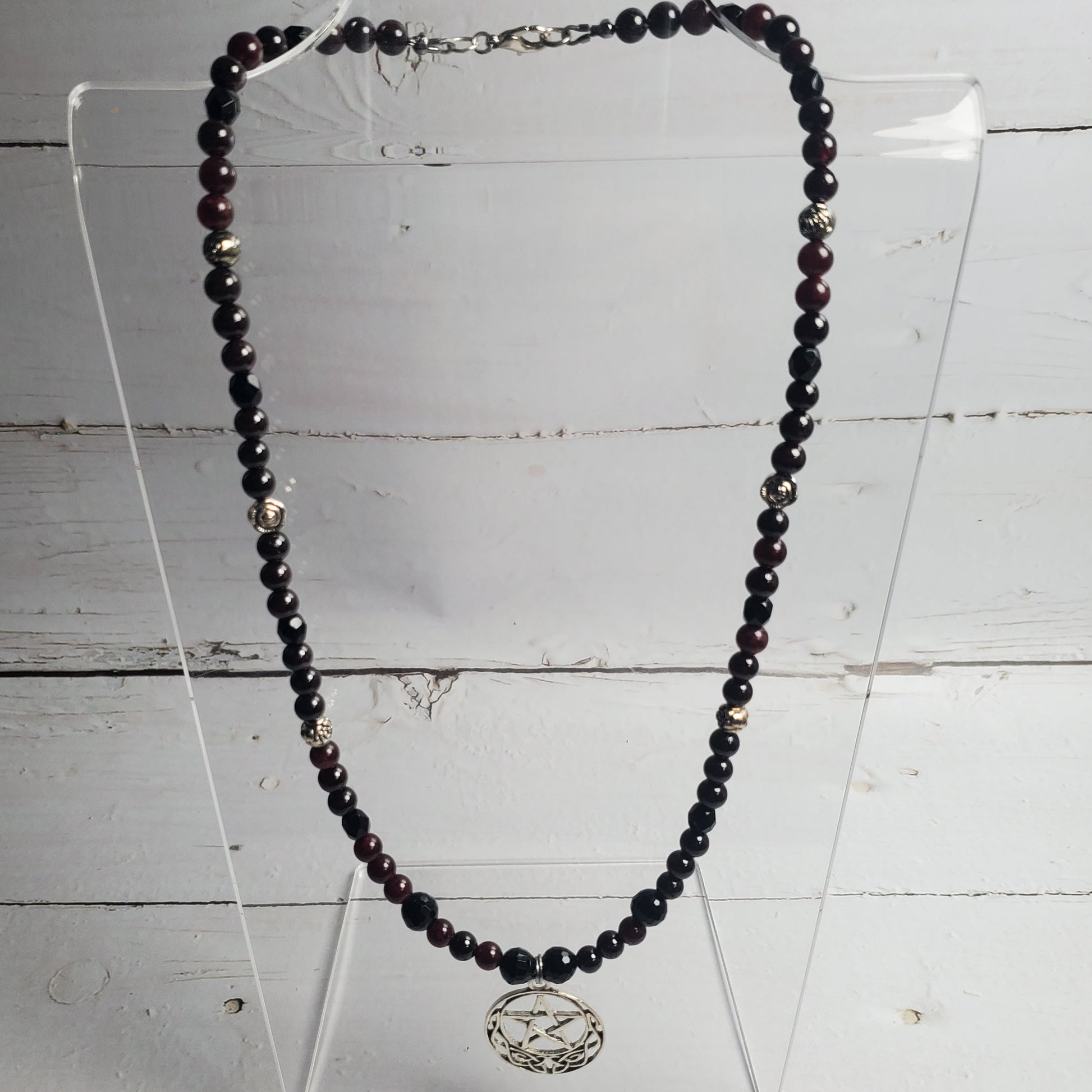 A beaded necklace with dark faceted beads and a silver-toned circular pendant, displayed on a clear acrylic stand against a white wood plank background.