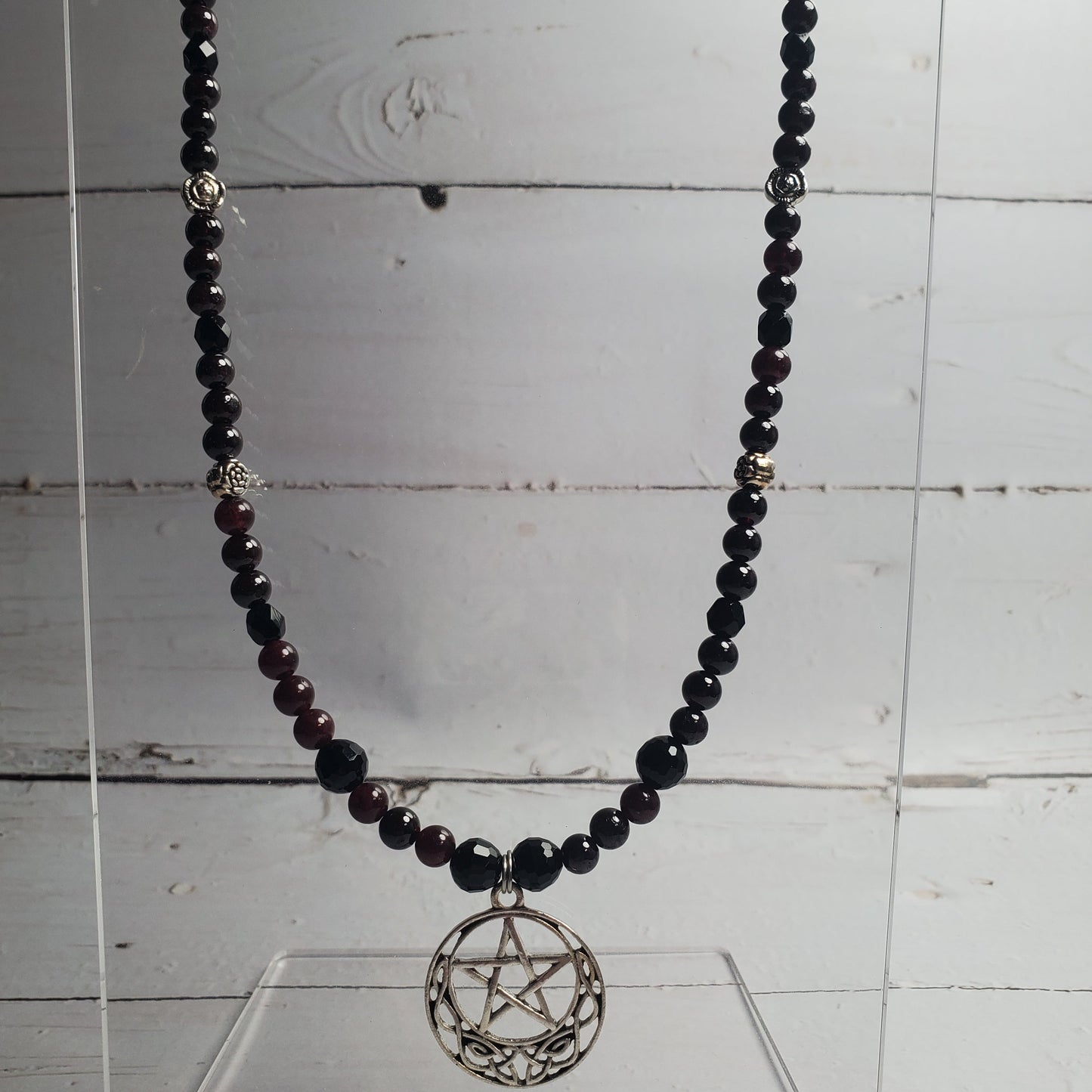 A beaded necklace with dark red and black faceted beads, featuring a silver-toned pentagram pendant, displayed on a clear acrylic stand against a white wood plank background.
