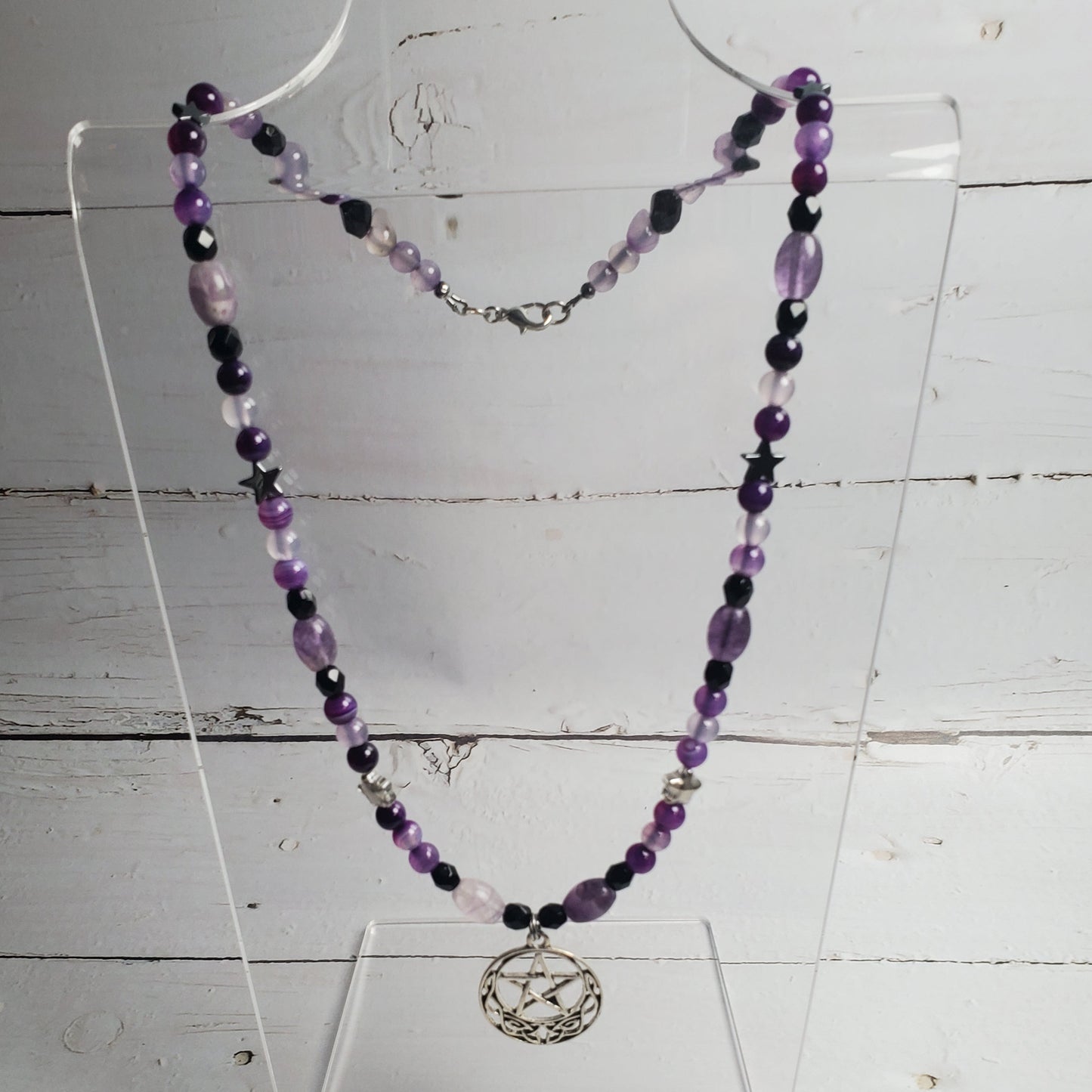 A beaded necklace with purple, black, and light pink beads, featuring a silver-toned circular pendant with a bird design, displayed on a clear acrylic stand against a white wood plank background.