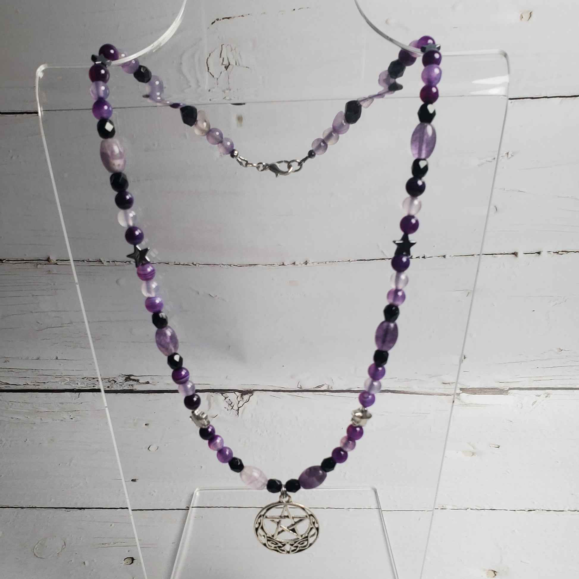 A beaded necklace with purple, black, and light pink beads, featuring a silver-toned circular pendant with a bird design, displayed on a clear acrylic stand against a white wood plank background.