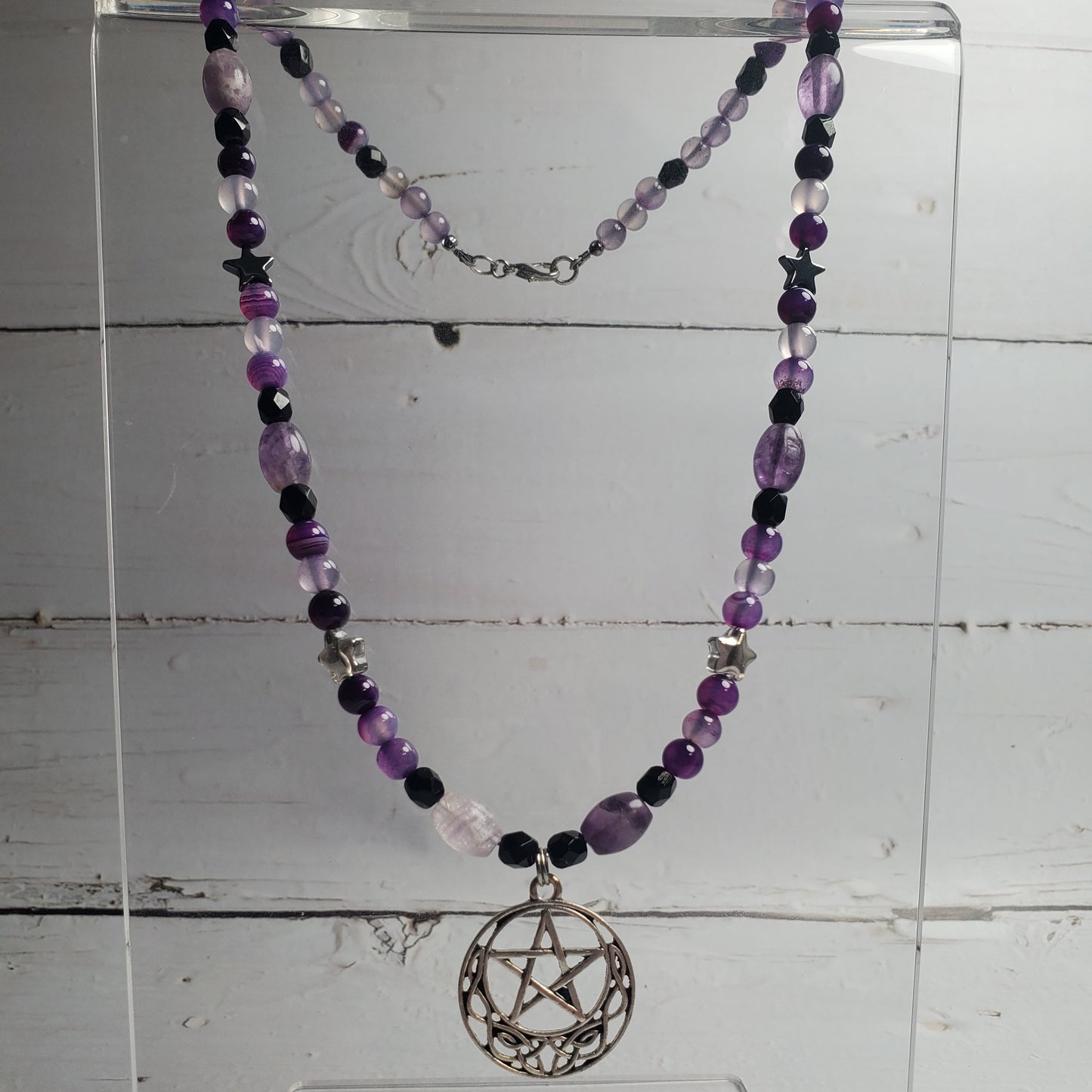 A beaded necklace with purple and black beads, star charms, and a silver pentagram pendant, displayed on a clear acrylic stand against a white wood plank background.