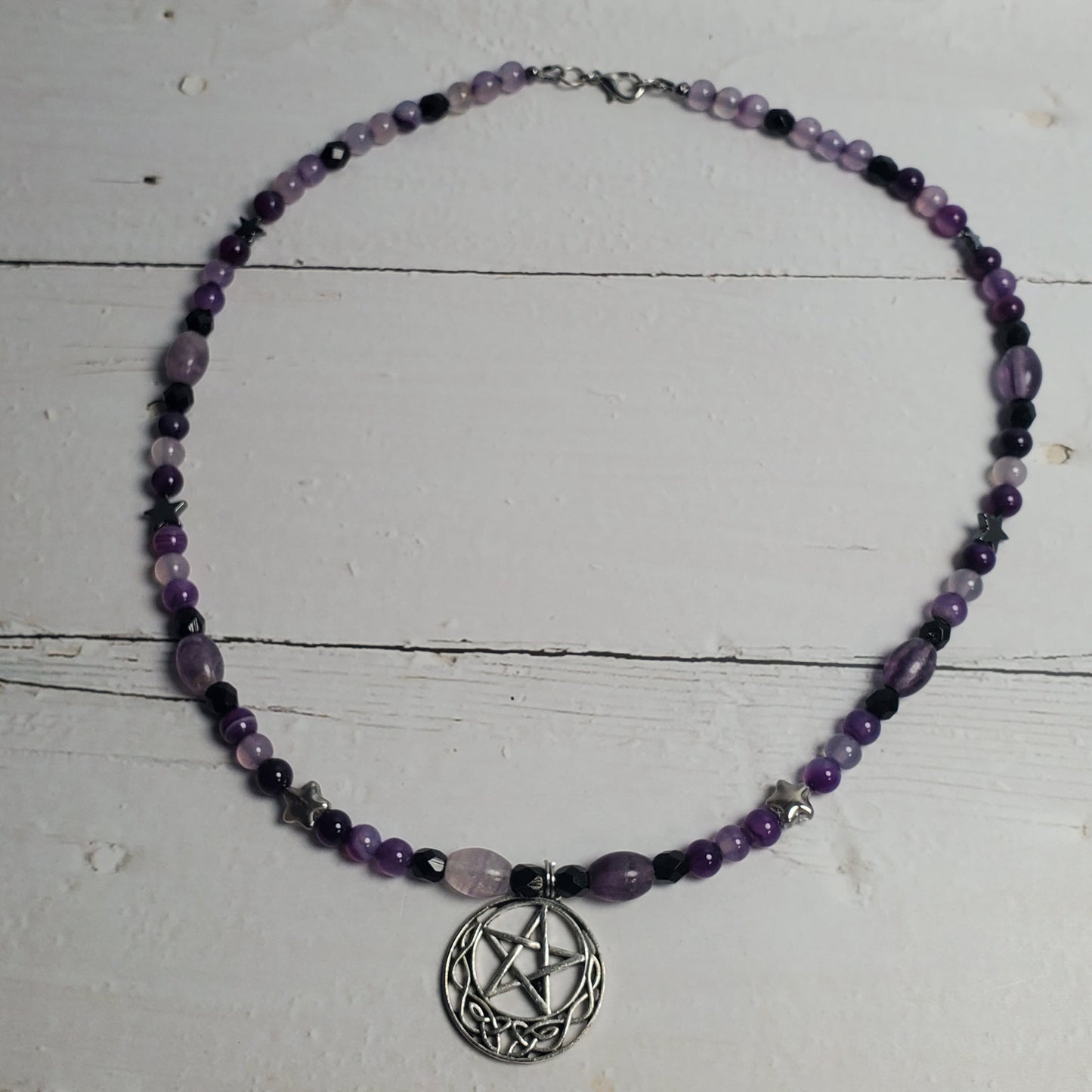 A beaded necklace with purple and black beads, featuring a central silver-toned pentagram pendant, displayed on a distressed white wooden surface.