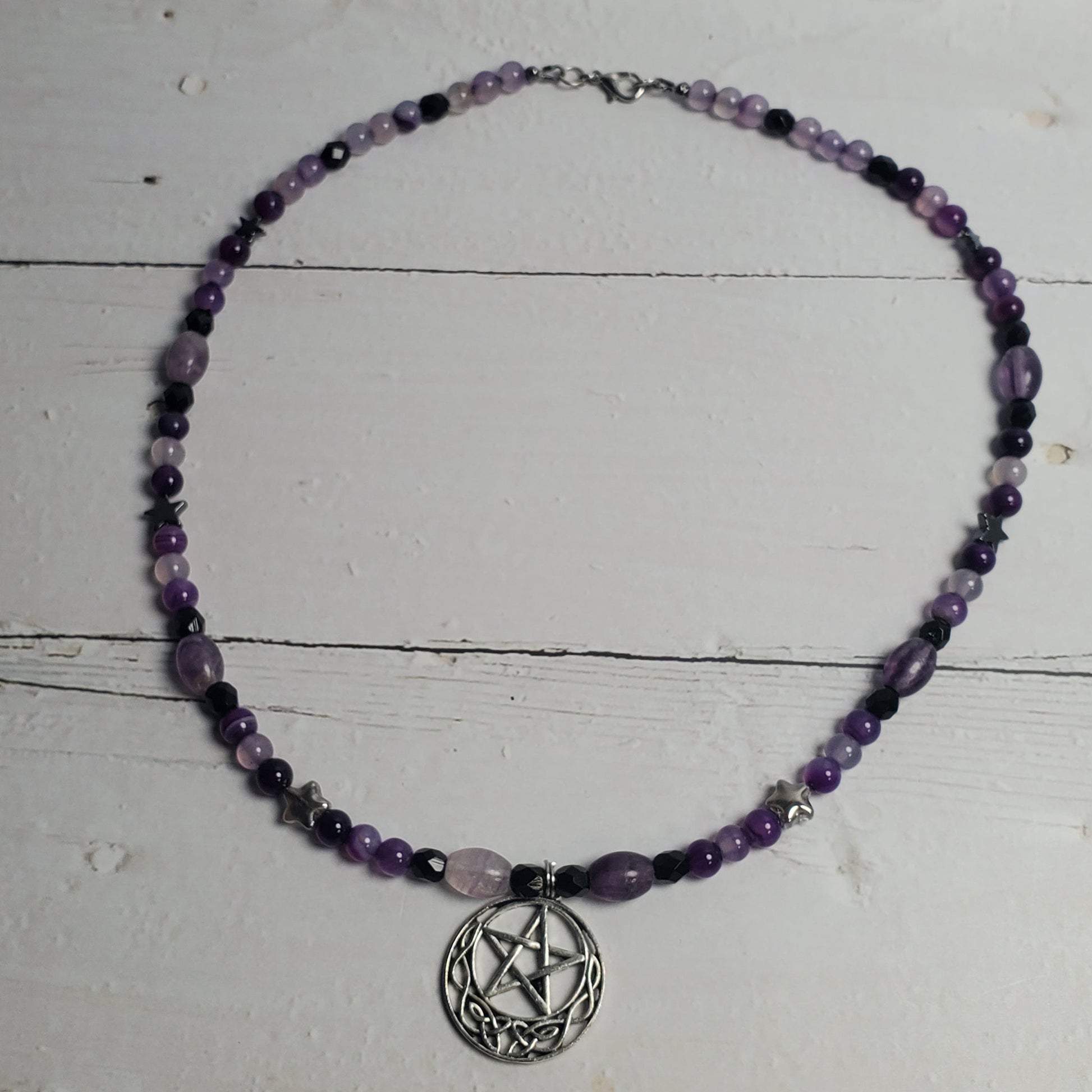 A beaded necklace with purple and black beads, featuring a central silver-toned pentagram pendant, displayed on a distressed white wooden surface.