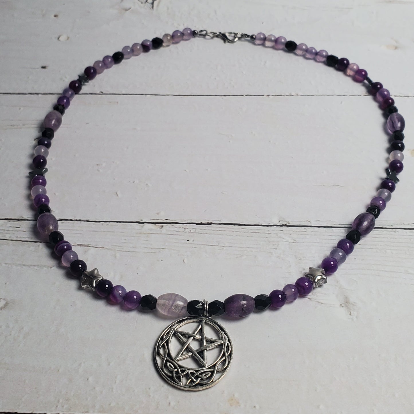 A beaded necklace with purple and black beads, featuring a silver pentagram pendant, displayed on a white wooden surface.
