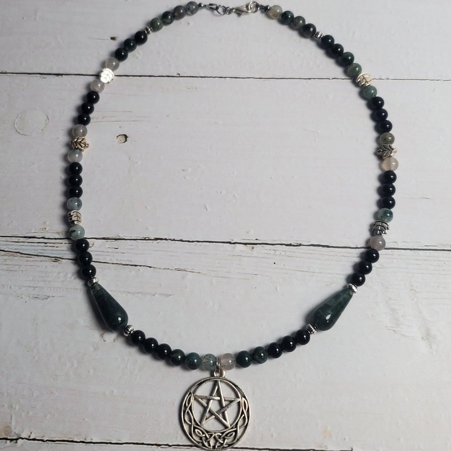 A beaded necklace featuring dark green and black beads, a silver pentagram pendant, and a large dark green teardrop bead, displayed on a white wooden background.