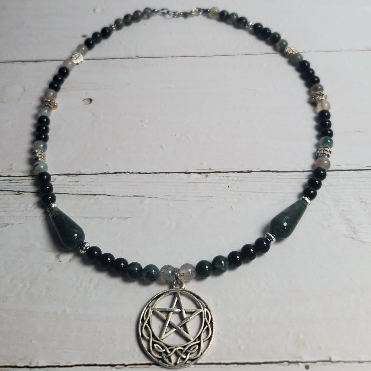 A beaded necklace featuring dark green and black beads, silver-toned spacer beads, and a central silver-toned pentagram pendant, displayed on a white wooden surface.