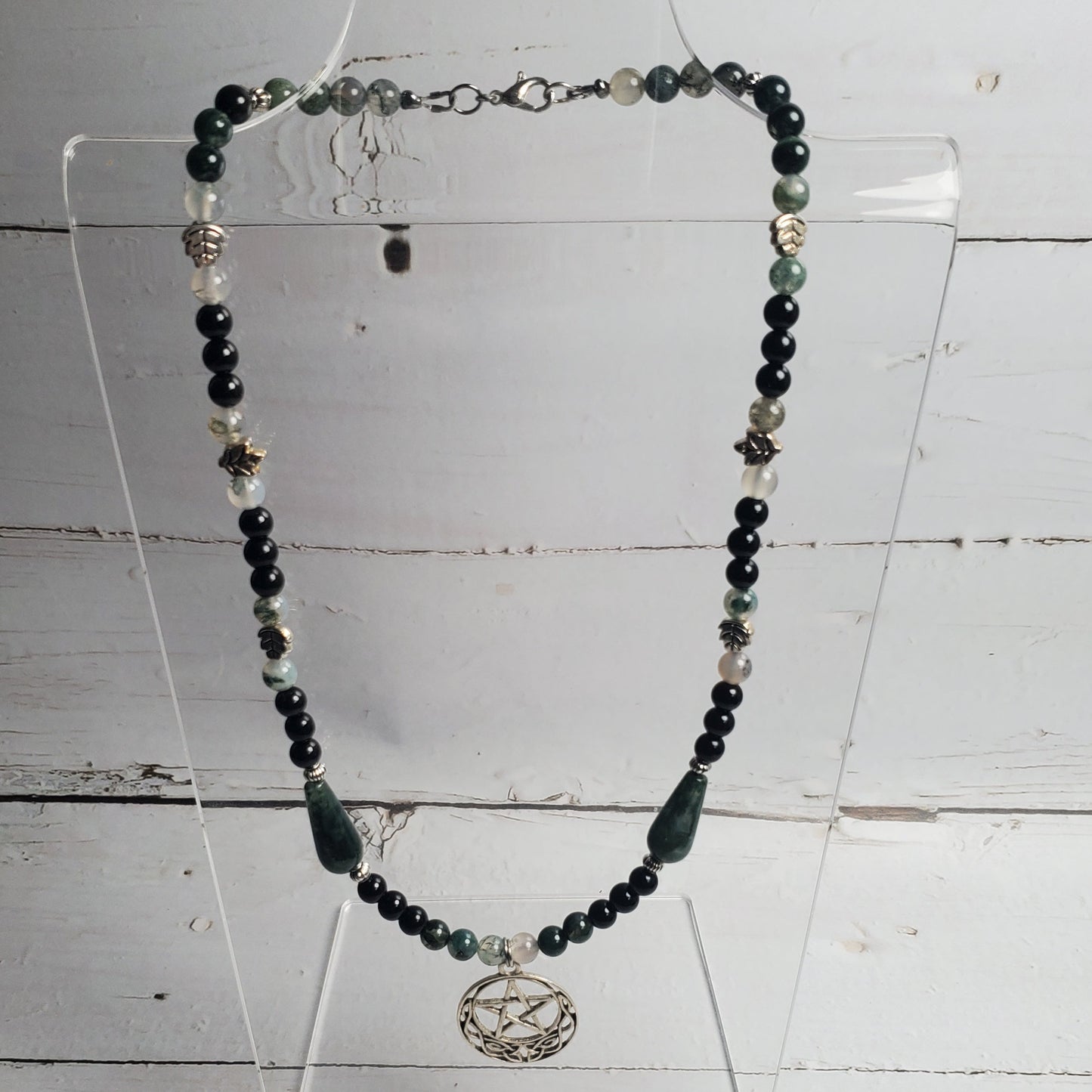 A beaded necklace displayed on a clear acrylic stand against a white wood plank background. The necklace features dark black beads, green stone beads, small silver spacer beads, and a central silver-toned circular pendant with a bird design.