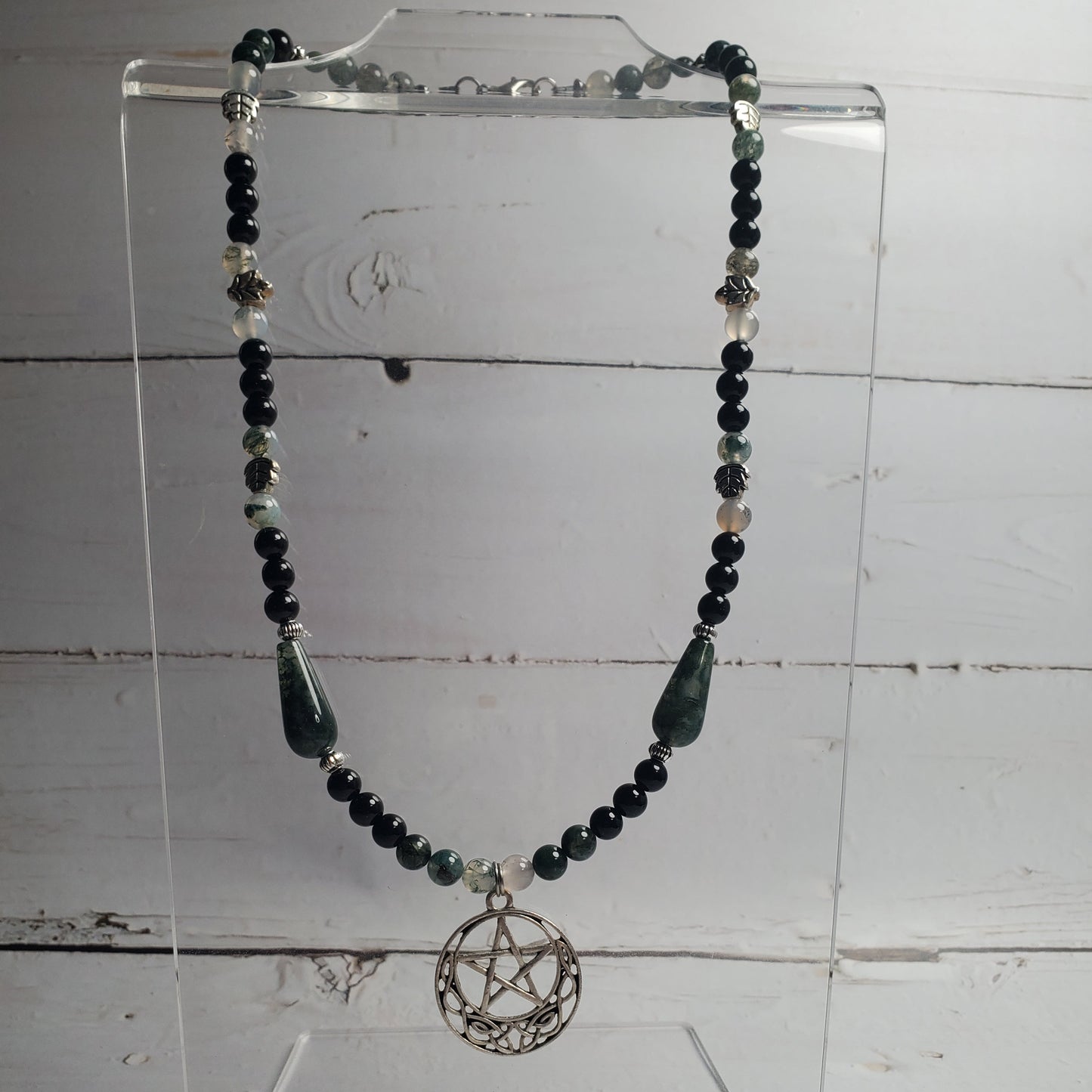 A beaded necklace with dark green and black beads, silver-toned spacer beads, and a silver pentagram pendant, displayed on a clear acrylic stand against a white wood plank background.