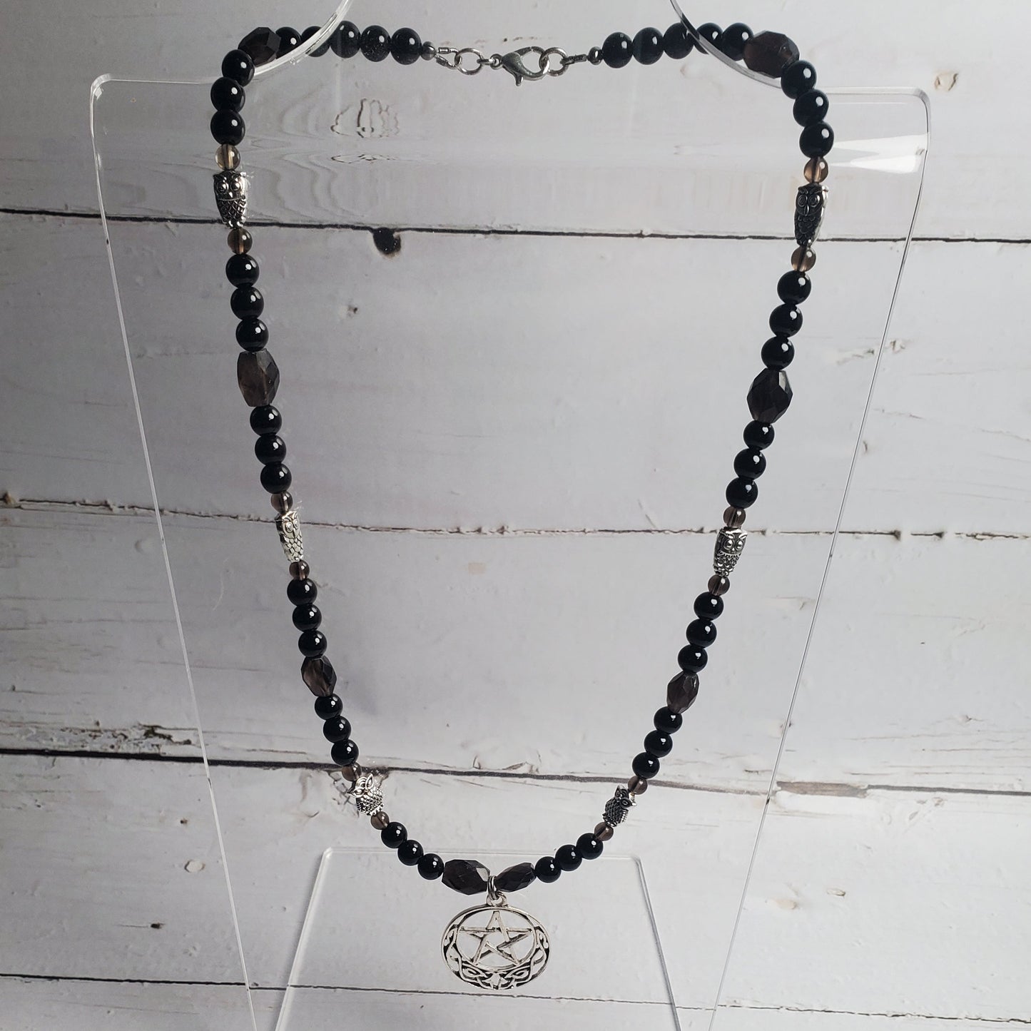 A beaded necklace with dark black and gray beads, silver spacer beads, and a silver-toned circular pendant, displayed on a clear acrylic stand against a white wood plank background.